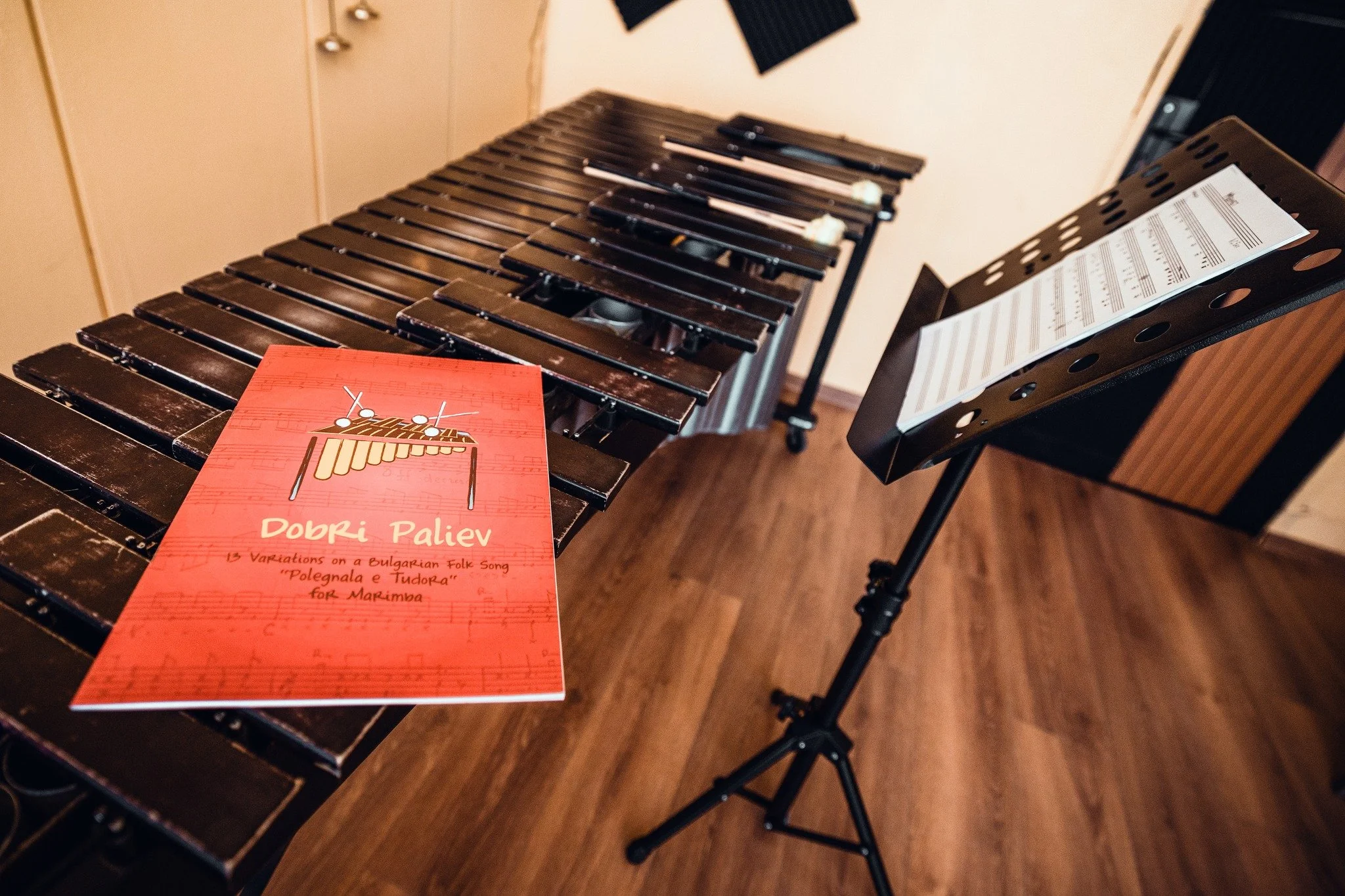 A music stand with sheet music and a red booklet titled 'Dobrí Paliev' placed on a wooden xylophone, with black soundproof panels on the wall in the background.