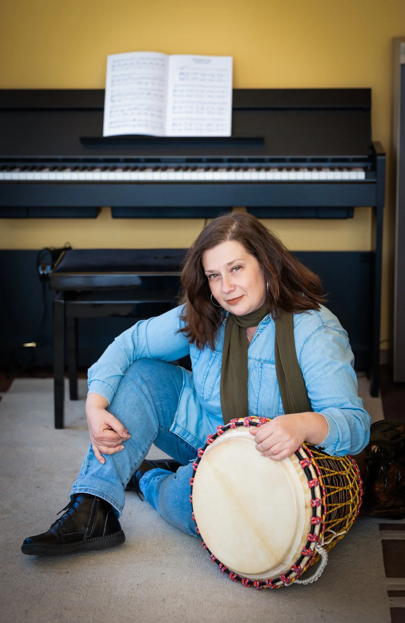 A woman with long brown hair sitting on the floor, playing a large drum with a drumstick, in front of a person lying on the floor. There is a black piano with an open music book on it behind her.