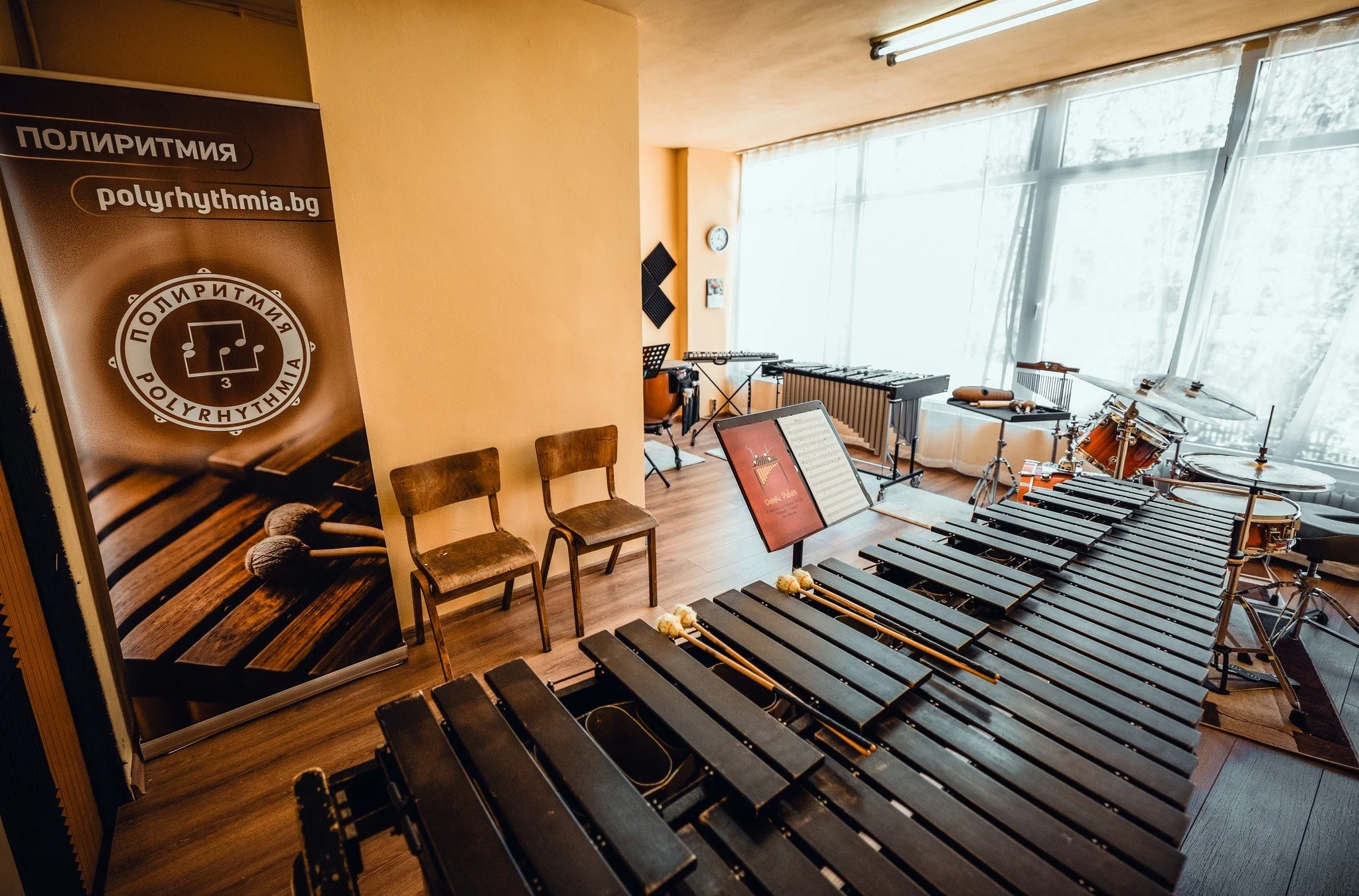 A music classroom with percussion instruments, including a large xylophone, drums, and maracas, set up for a lesson. There are chairs, a music stand with sheet music, and large windows letting in natural light. A banner with the logo and website for 