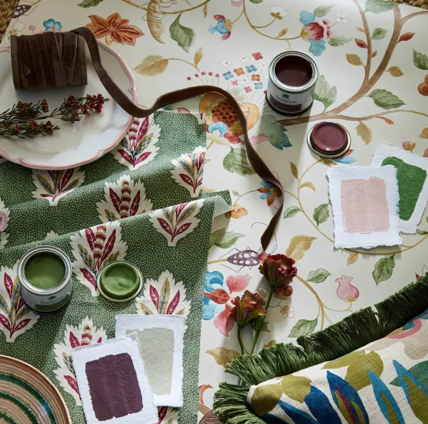 Various fabric swatches, jars of fabric dye, a paintbrush, and flowers on a floral-patterned tablecloth, suggesting fabric dyeing or crafting activities.