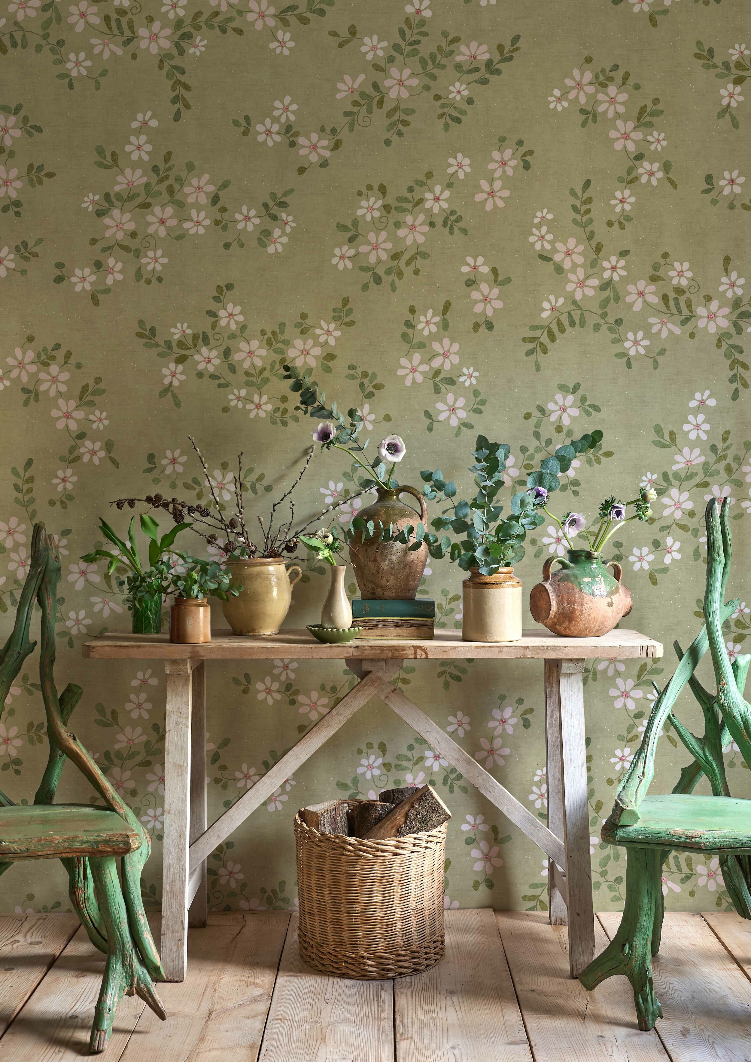 A rustic wooden table with various vases and plants, set against a floral wallpapered wall, flanked by two distressed green chairs.