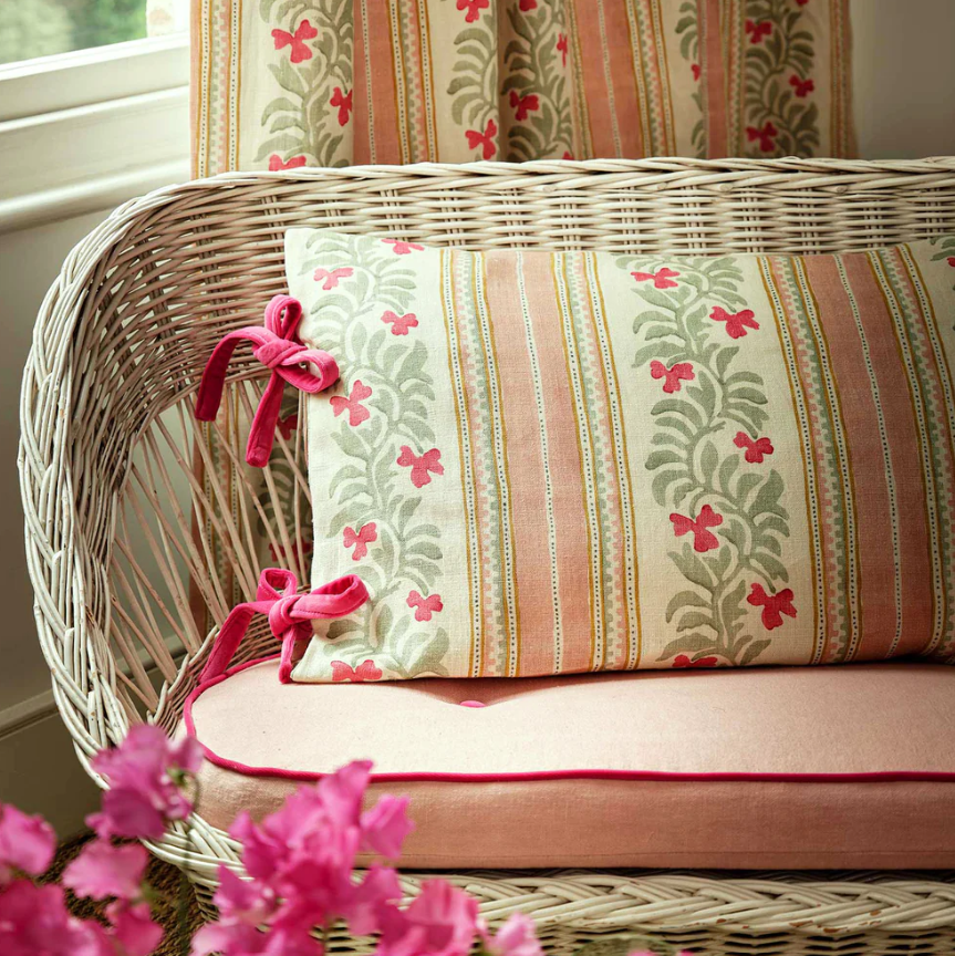 A wicker bench with a floral cushion and pillow, decorated with pink bows, placed near a window with floral curtains.