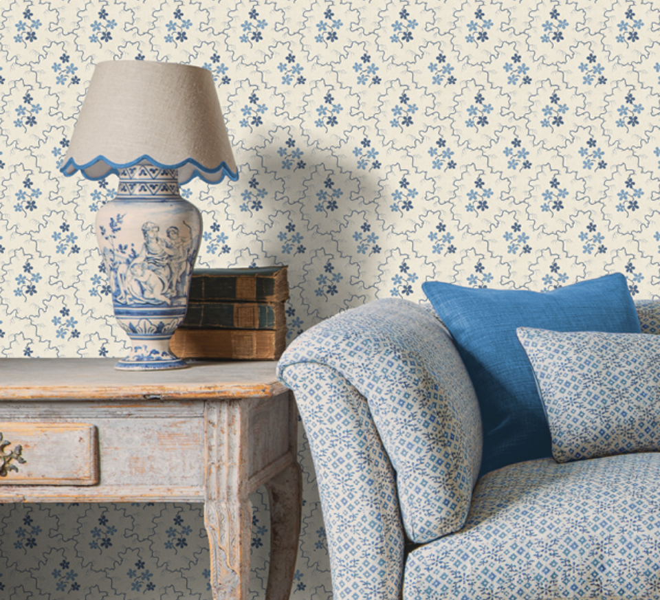 A vintage living room corner with a distressed white side table, a blue and white ceramic lamp, a stack of old books, and a sofa with blue patterned pillows. The background features wallpaper with blue floral designs.