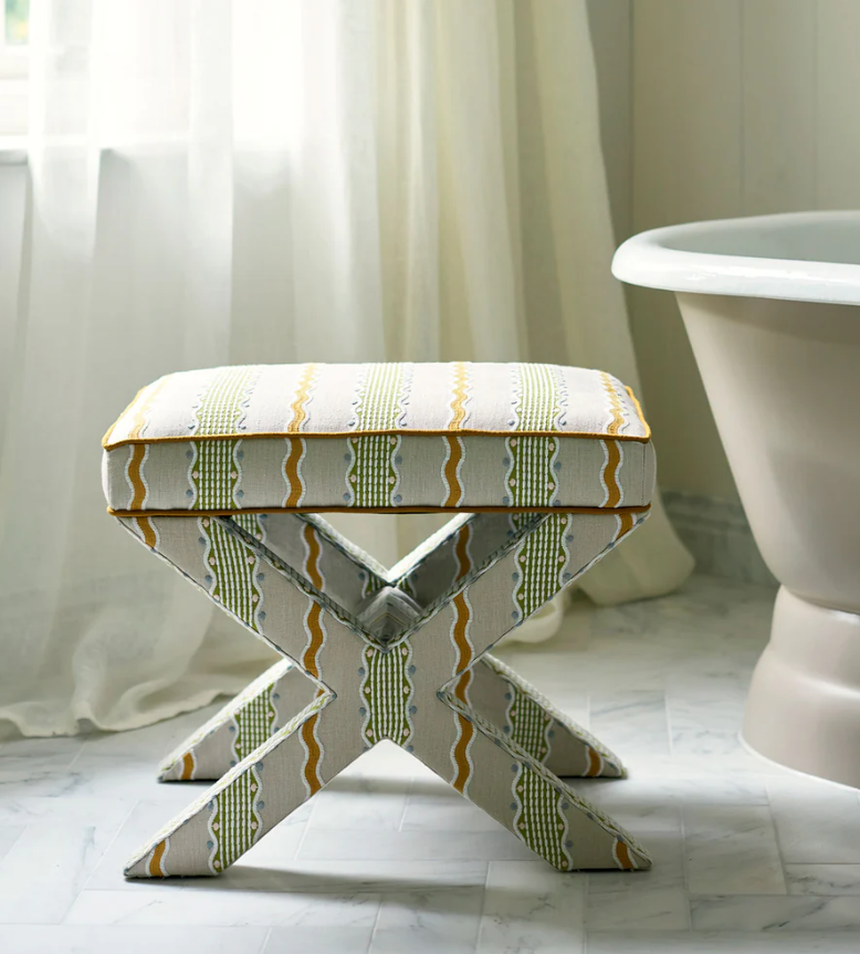 A cushioned folding stool with a patterned fabric cover, placed on a marble floor next to a white bathtub with a rounded edge, in a bathroom with sheer curtains and soft natural lighting.
