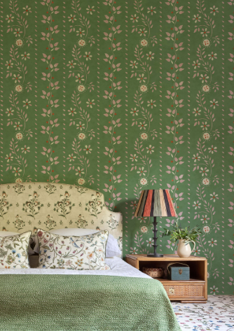 A bedroom with a green floral wallpaper, a patterned headboard, decorative pillows, a wooden nightstand with a potted plant, and a colorful striped table lamp.