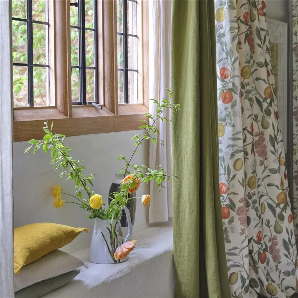 A window with wooden frame and glass panes, with various curtains including a patterned curtain with fruits, a solid green curtain, and a white curtain. There is a white ledge with a yellow pillow and a white pitcher holding a bouquet of flowers with pink and yellow blooms.