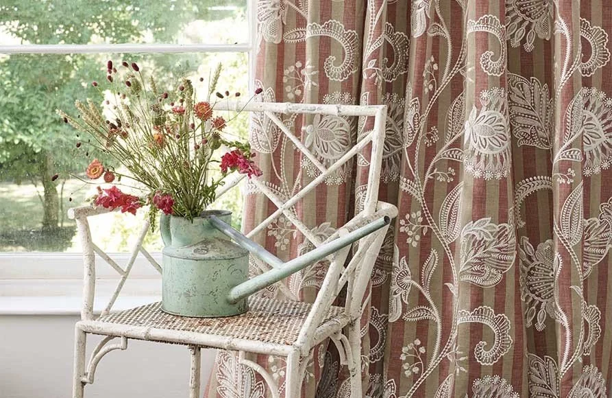 A framed, white metal chair with a can of flowers on the seat, sitting beside a window with lace curtains.