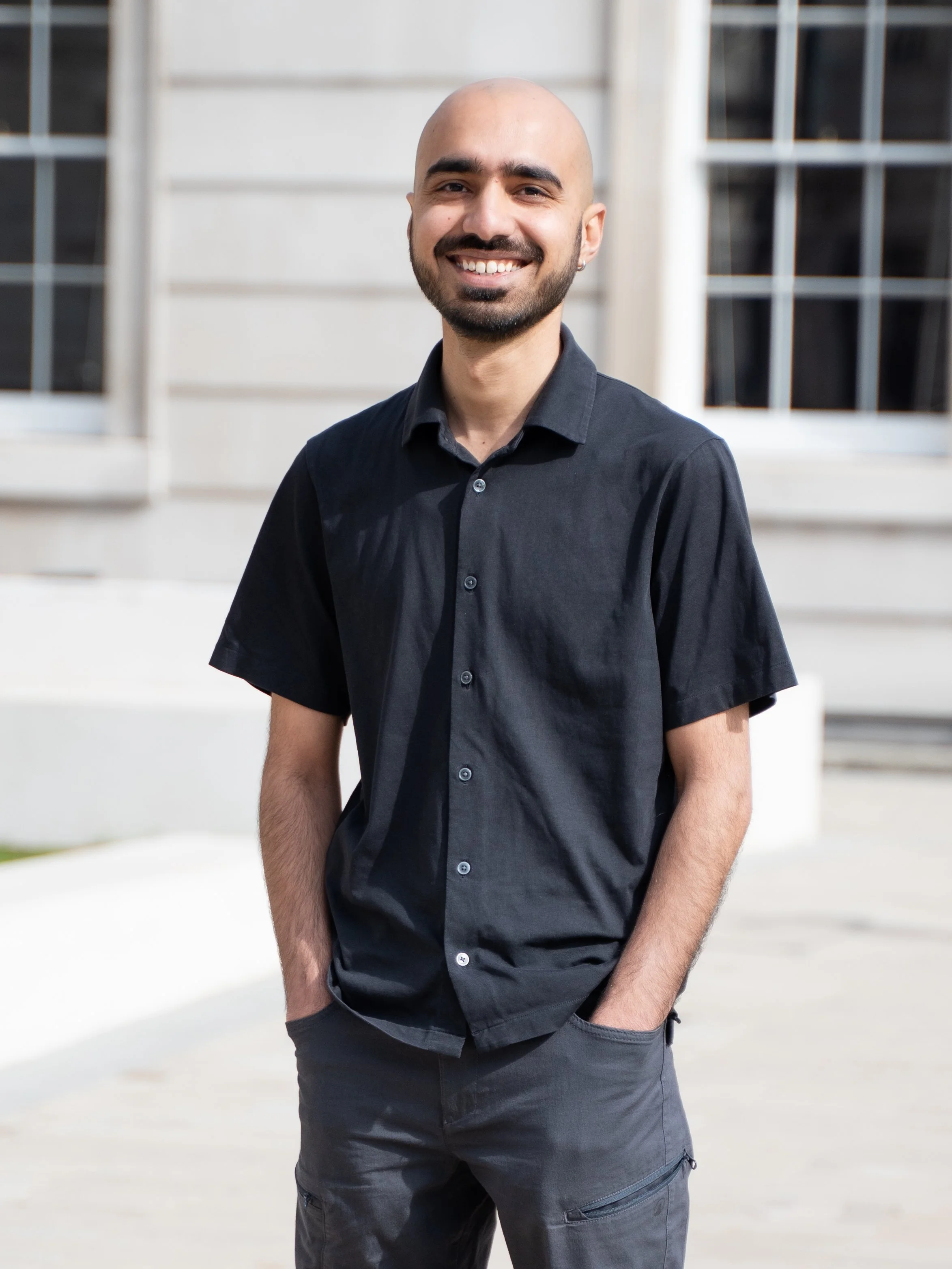 A smiling man with a shaved head, beard, and earring wearing a black short-sleeved button-up shirt and dark pants, standing outside in front of a white building with windows.