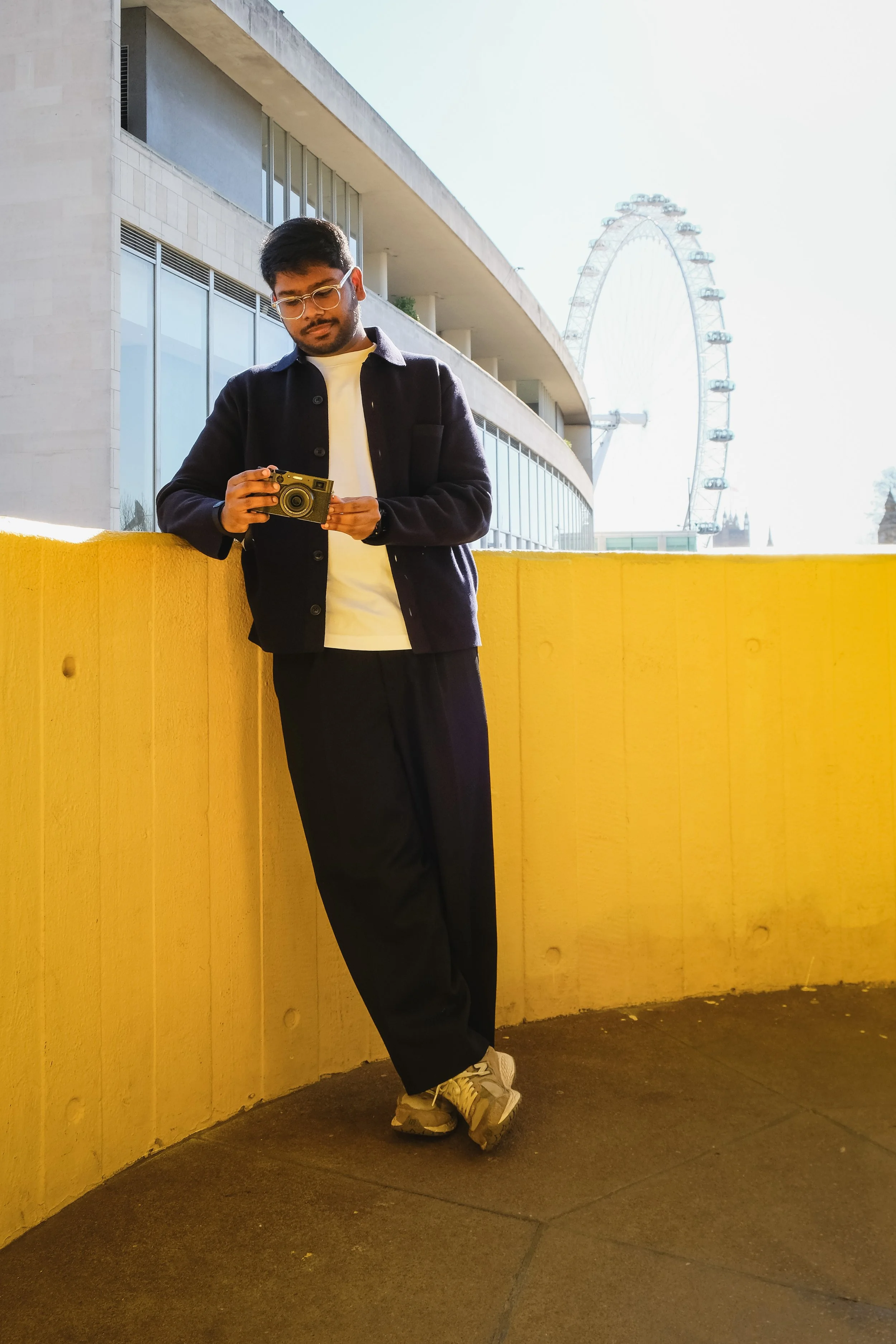 A young man with glasses, wearing a black jacket, white T-shirt, black pants, and sneakers, leaning against a yellow wall while looking at a vintage camera in his hands. In the background, there is a large Ferris wheel and modern building with windows.