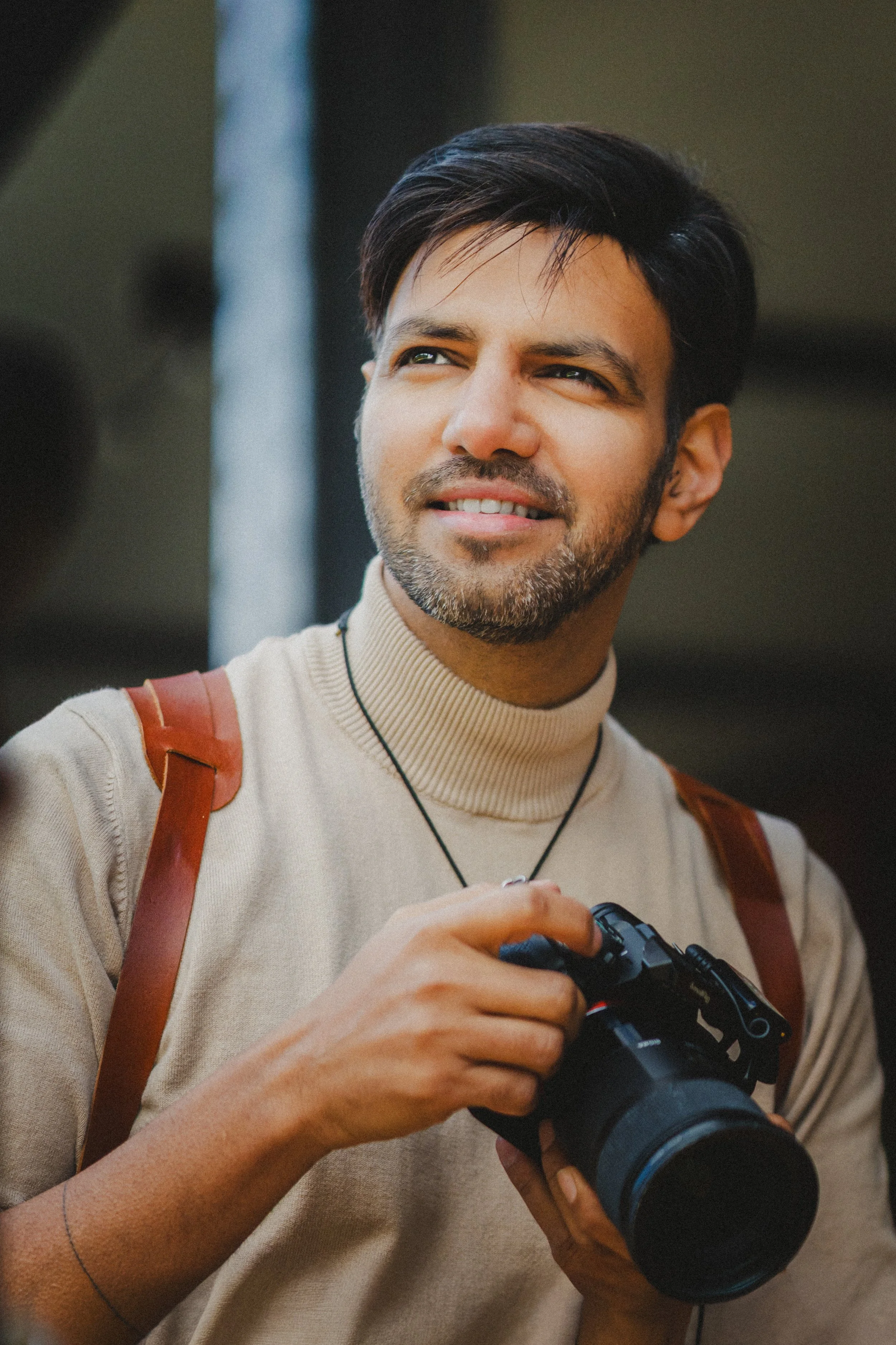 A man with dark hair, a beard, and light skin holding a camera, wearing a beige sweater, a necklace, and a brown strap over his shoulder, outdoors.
