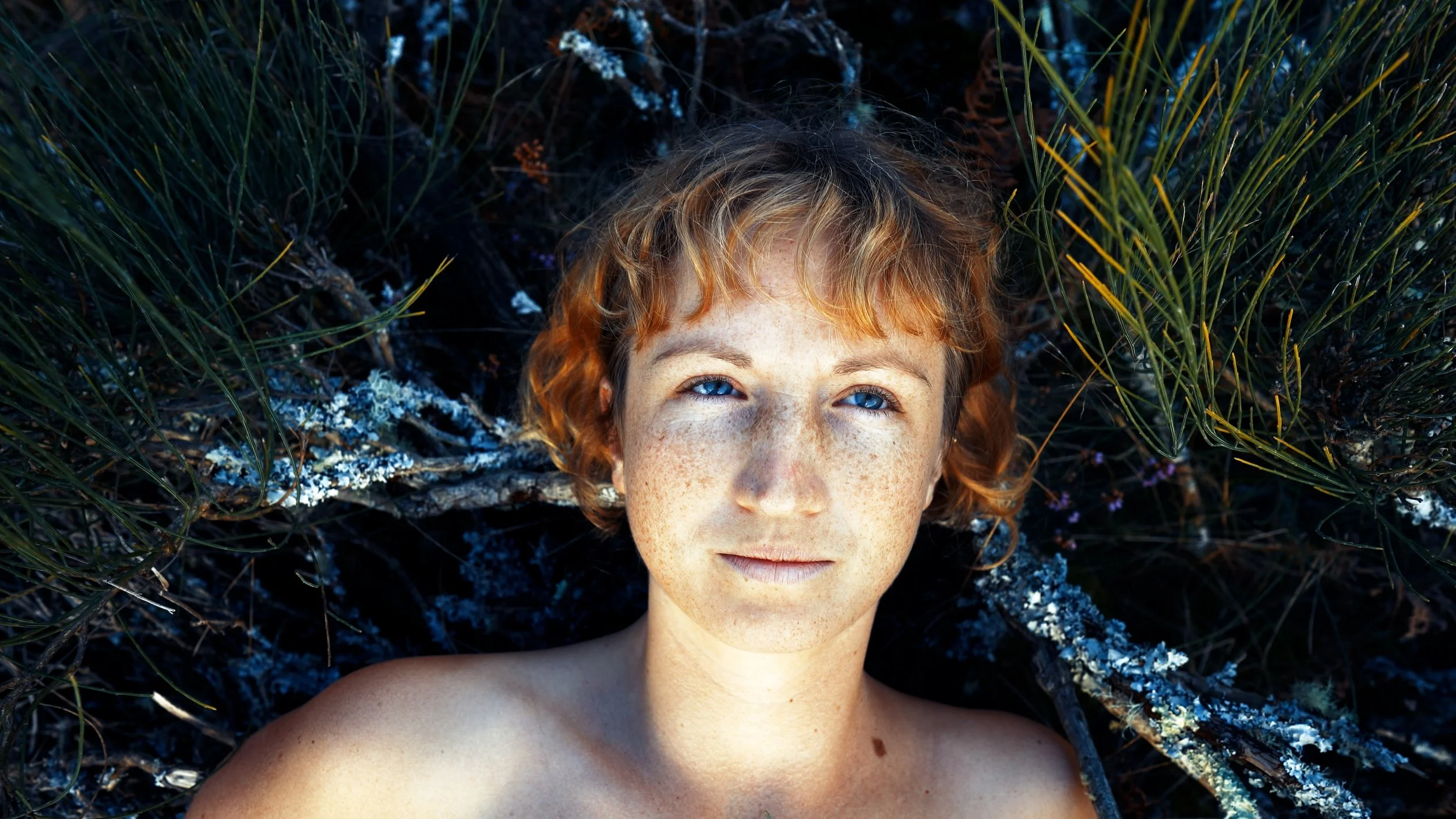 Close-up of a young woman with red hair and freckles lying on the ground surrounded by green grass and branches.