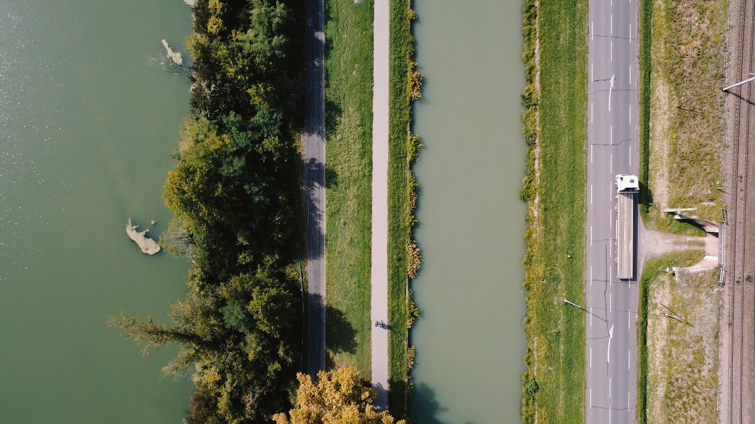 An aerial view of a city canal with a small white boat docked along the sidewalk, greenery on both sides, and a nearby parking lot with a few cars.