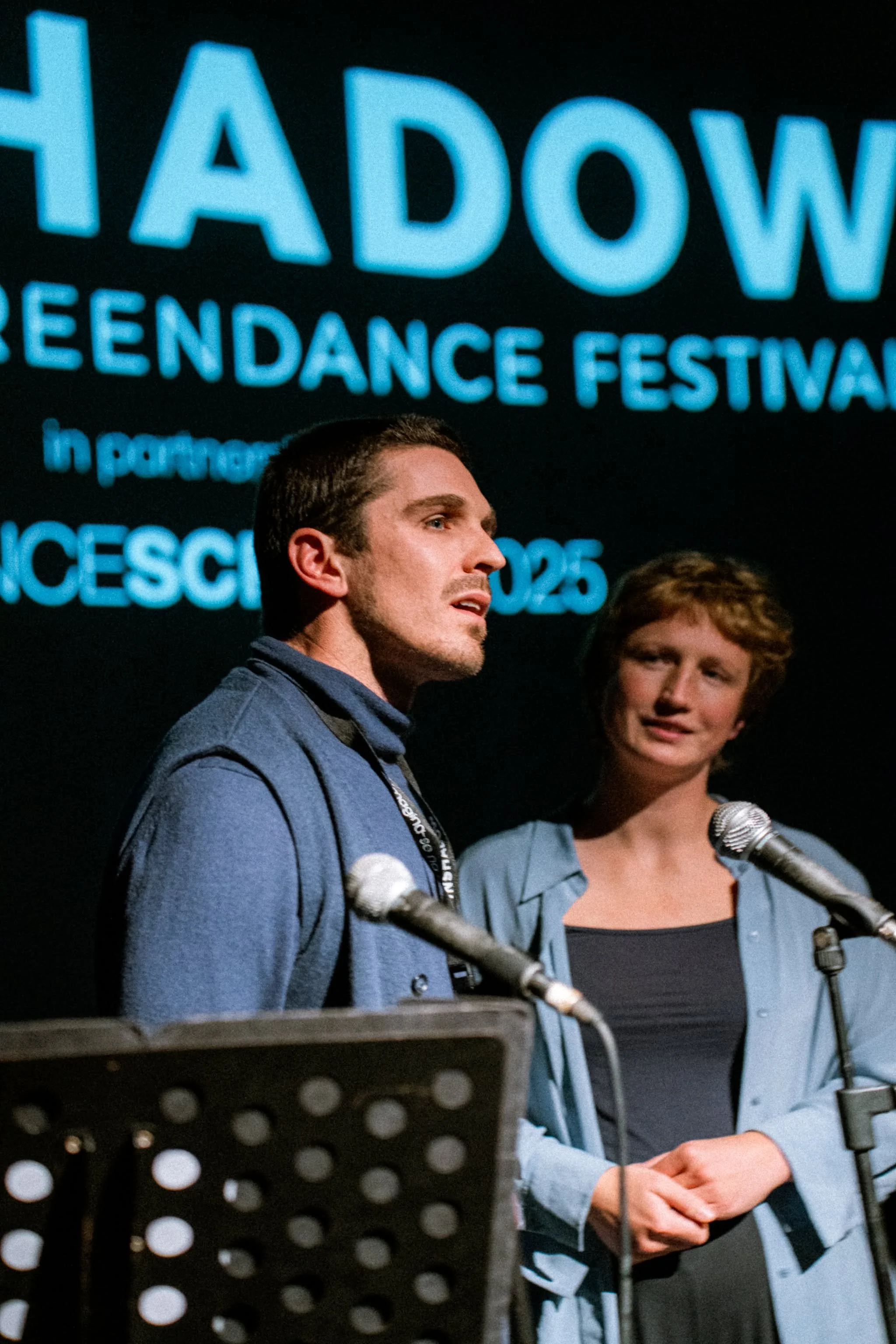 Two people standing at microphones during a panel or presentation at the Shadow Green Dance Festival, with a large screen displaying the event's title and details behind them.