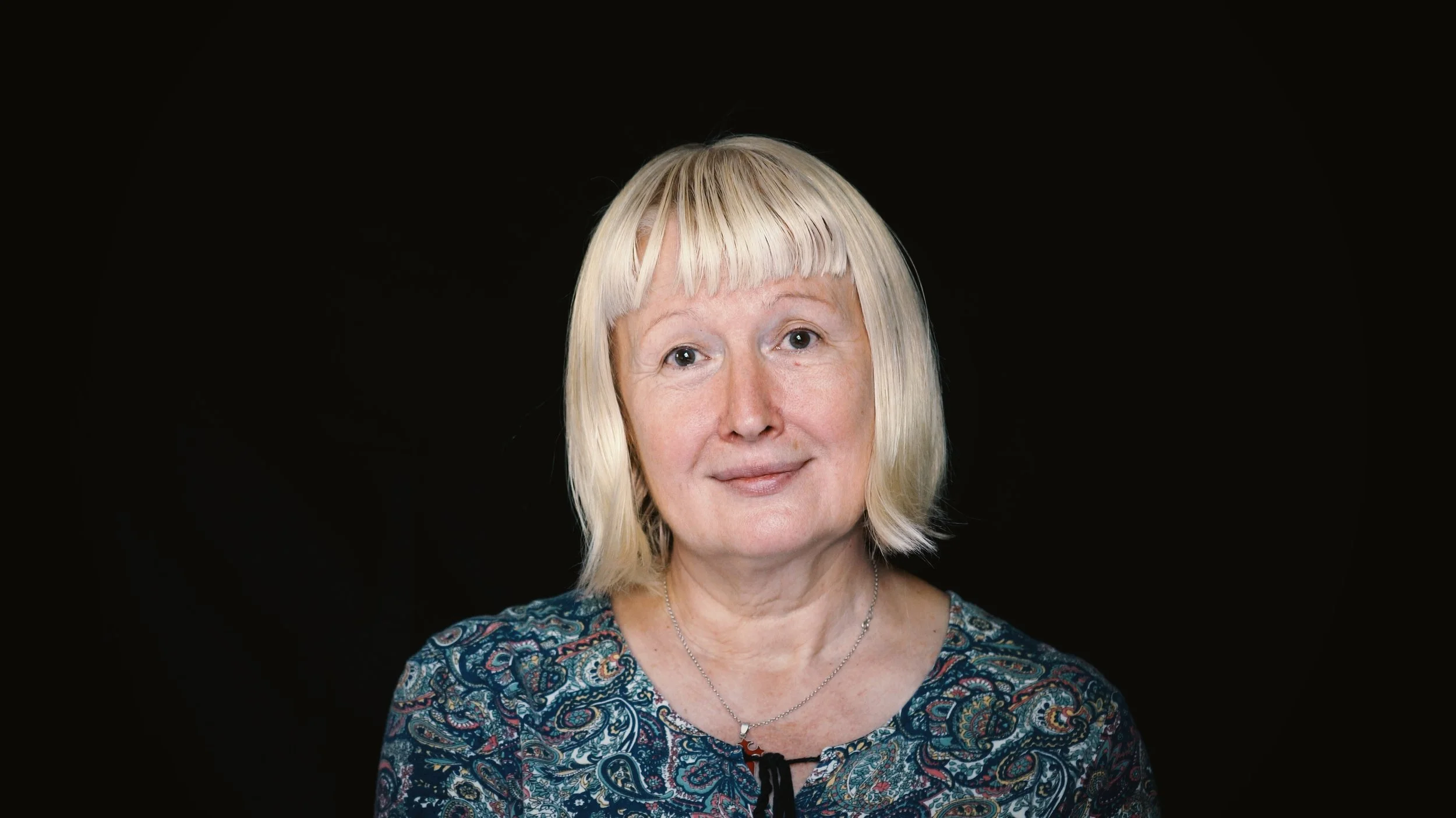 Close-up portrait of a middle-aged woman with short blonde hair, wearing a patterned blouse and a necklace, against a black background.