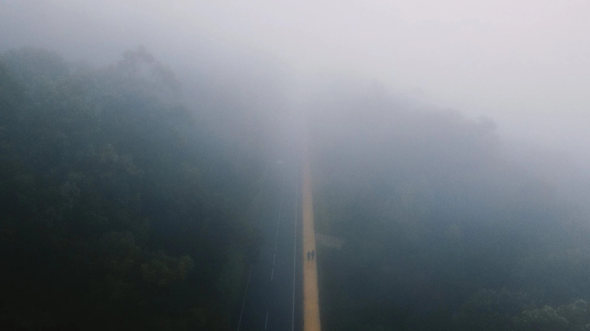 A lightning bolt strikes down through dark, stormy clouds over a rural area with trees and a road.