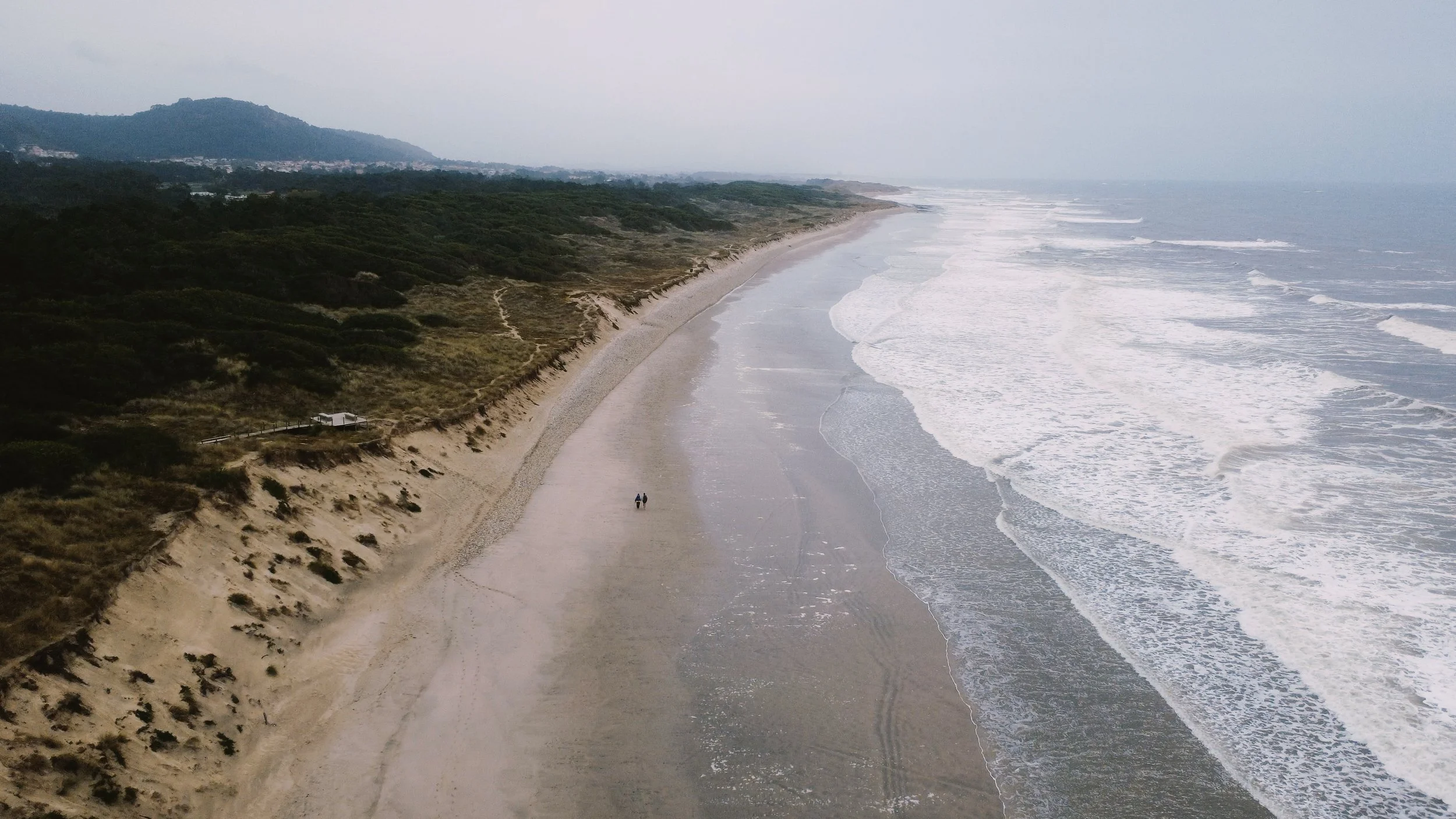 Aerial view of a beach with sand dunes and greenery on the left, waves on the right, and two people walking along the shoreline.