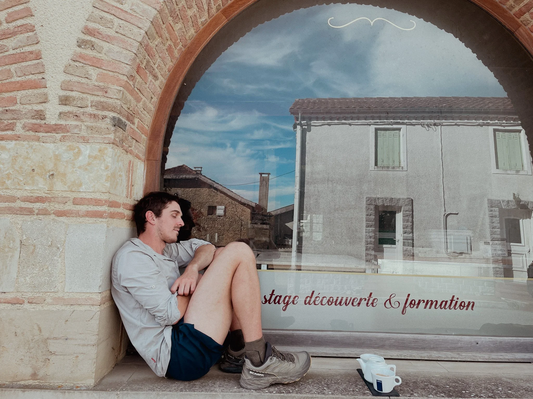 Young man sitting with his back against a brick wall, eyes closed, appearing relaxed, next to a glass window with a reflection of a building and sky outside.