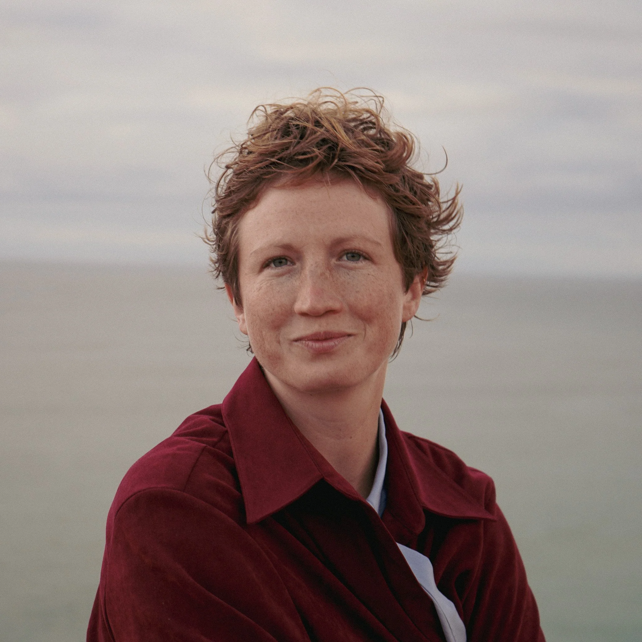 Close-up of a woman with short, curly red hair and blue eyes wearing a maroon jacket against a cloudy sky.