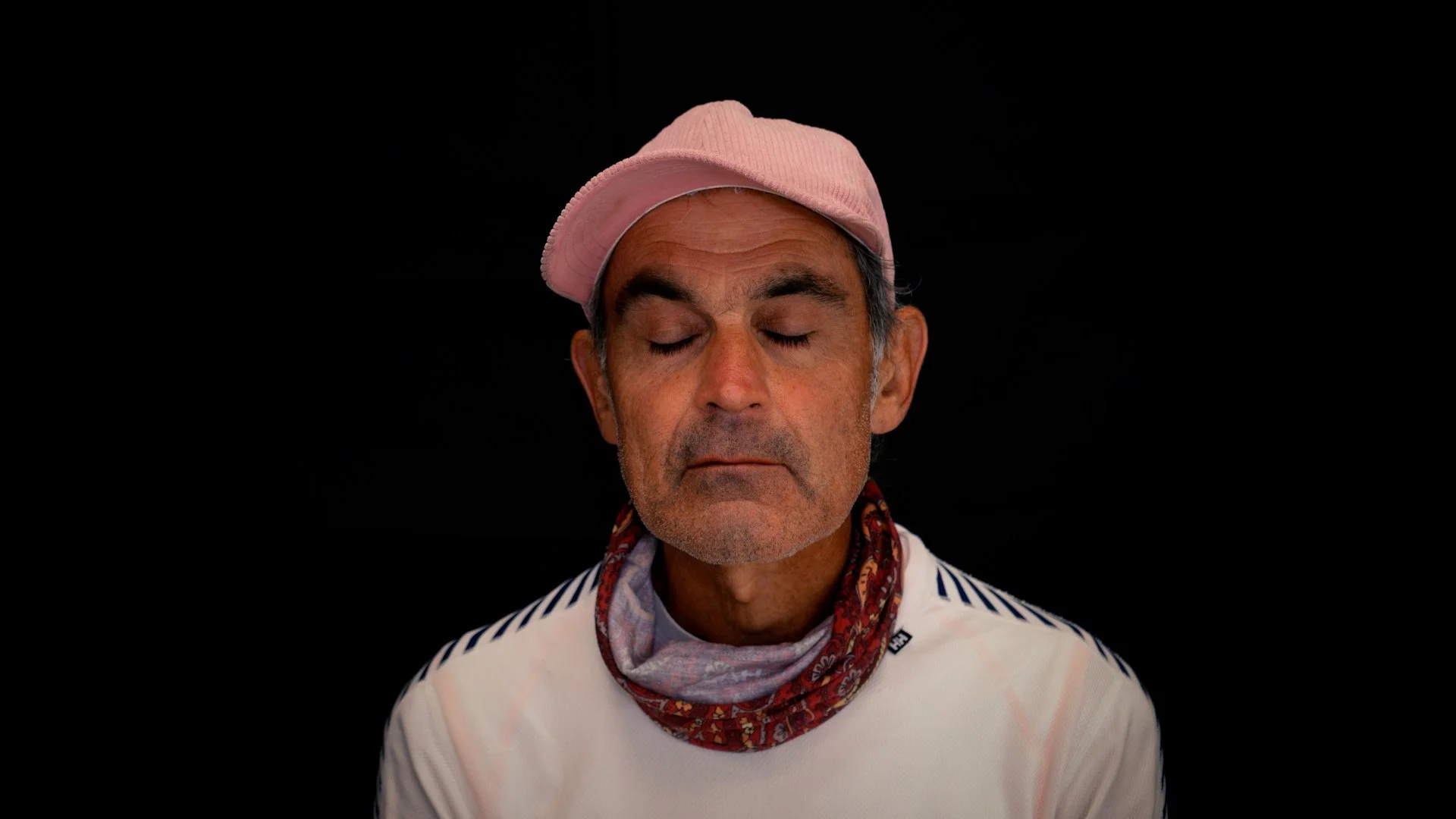 A middle-aged man with gray hair and stubble, eyes closed, wearing a pink cap, a patterned bandana around his neck, and a white striped shirt, against a black background.