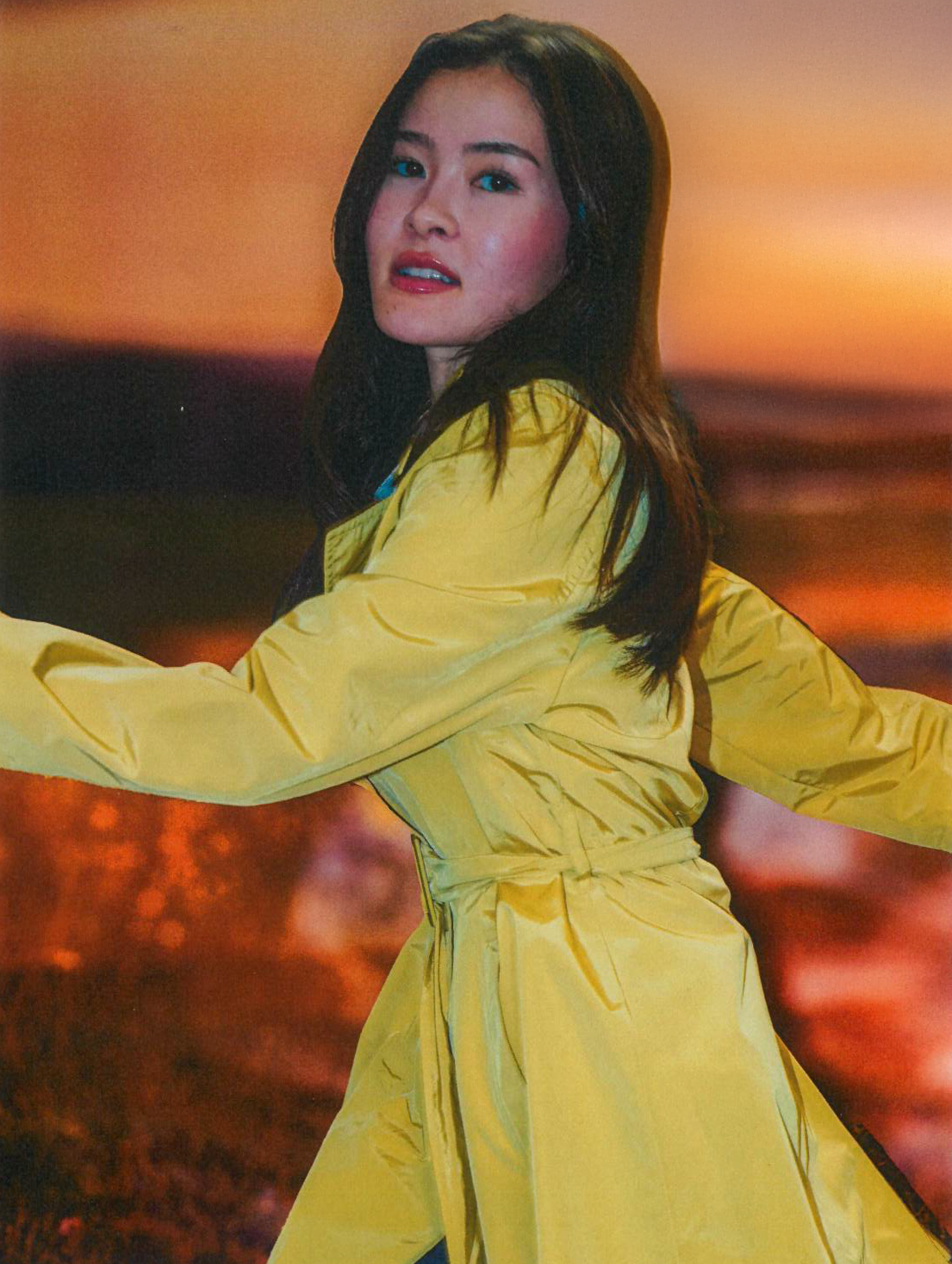 A woman with long dark hair wearing a yellow jumpsuit, standing outdoors at sunset with a blurred reddish sky background.