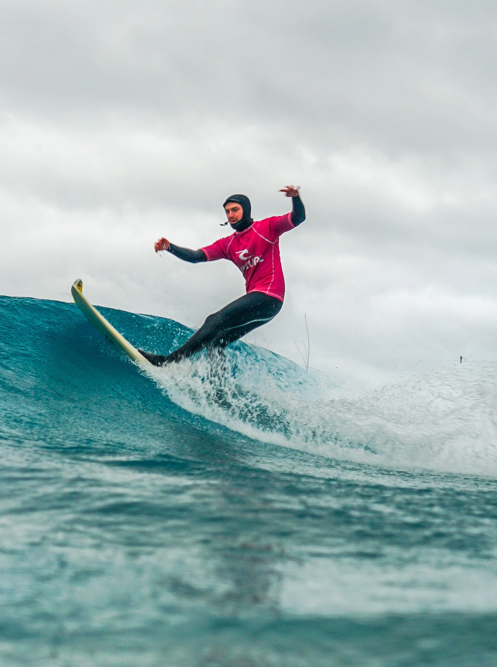 A person surfing on a wave in the ocean on a cloudy day, wearing a pink wetsuit and a black helmet.