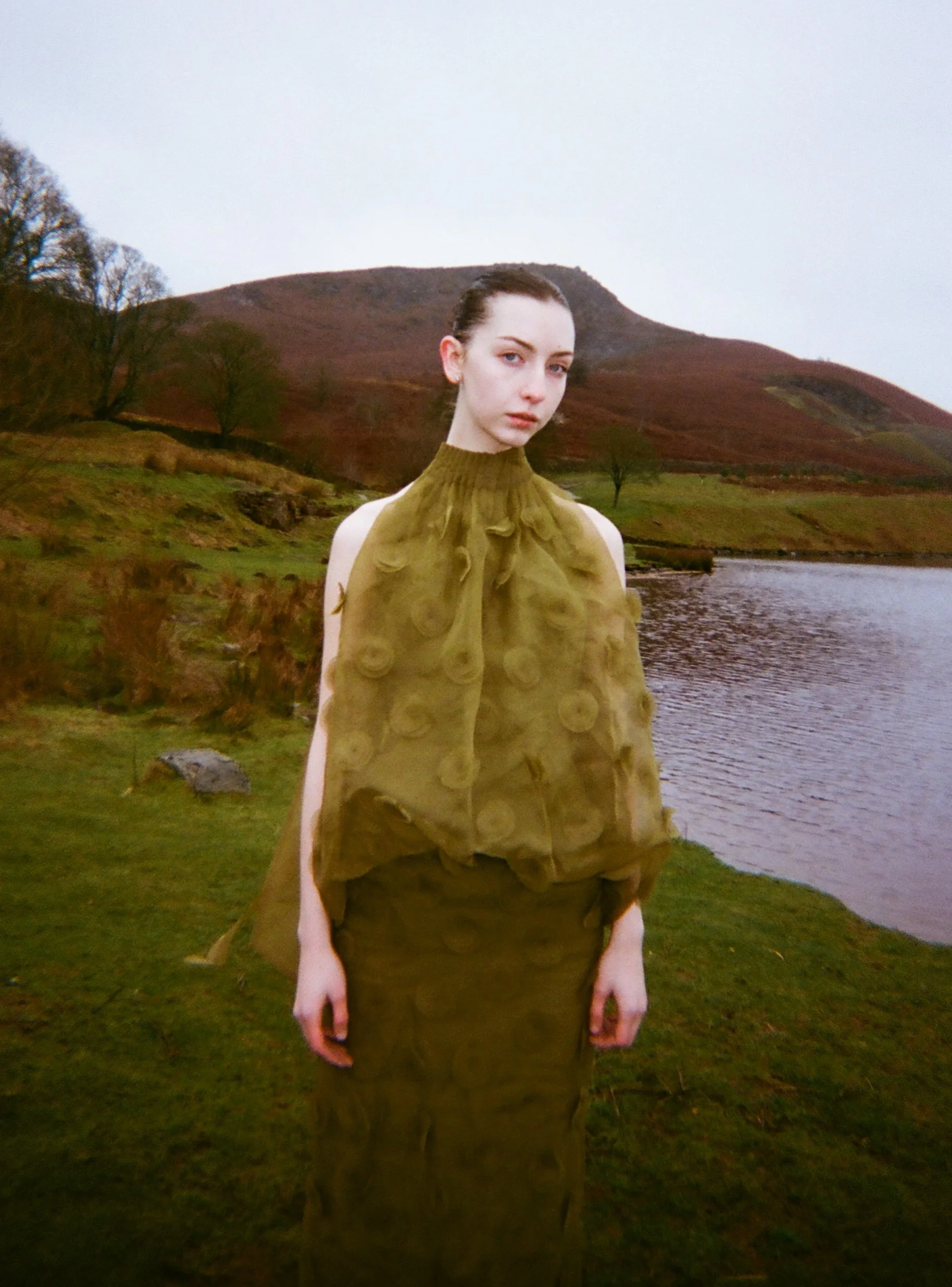 A woman standing outdoors near a body of water with hills in the background, wearing a patterned olive-green top and a long skirt.