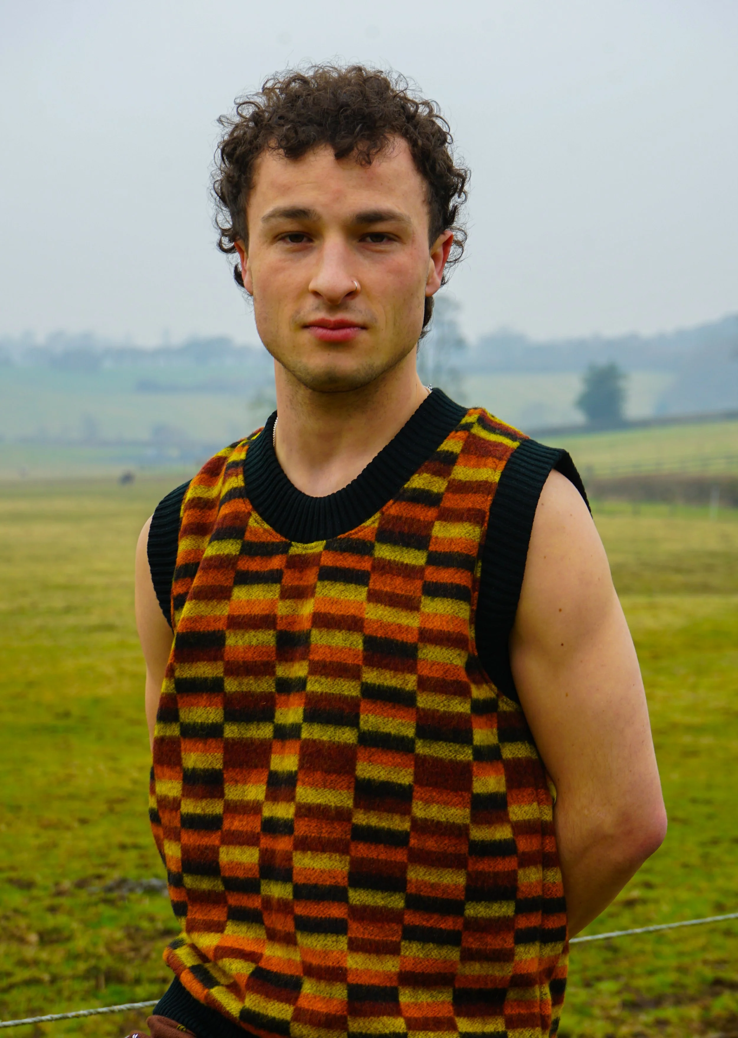 A young man with curly brown hair and a nose ring standing outdoors in a field with a cloudy, overcast sky.