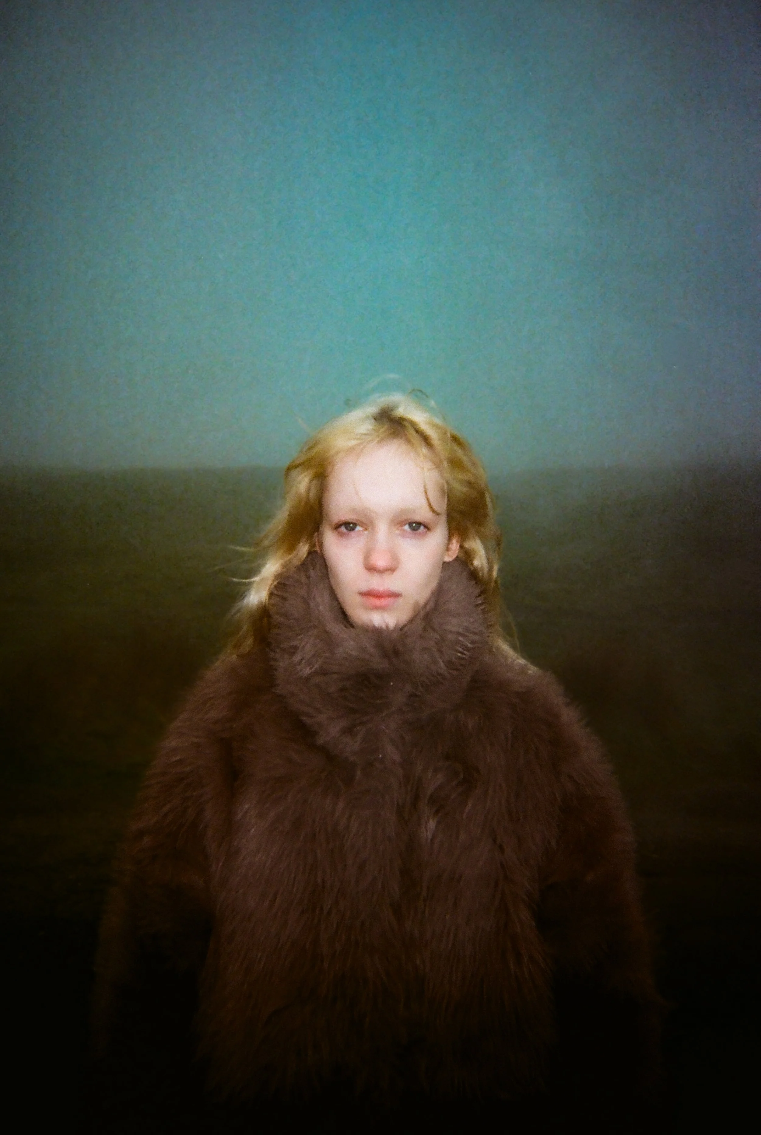 A young woman with blonde hair and fair skin wearing a brown fur coat, standing in front of a split-colored background with a gradient from teal at the top to dark brown at the bottom.