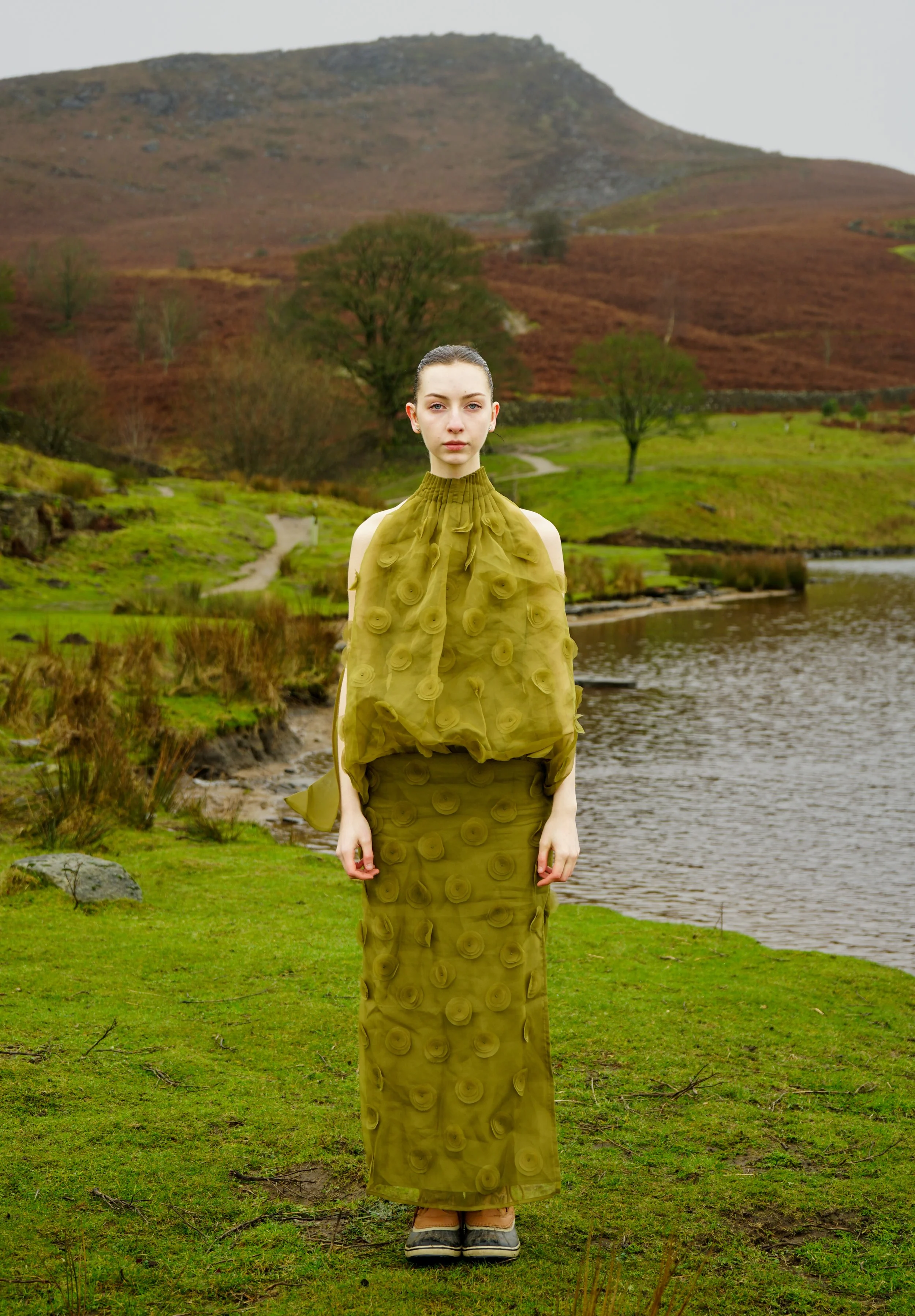 A woman standing outdoors near a river with hills and trees in the background, wearing a long green dress with circular patterns and closed-toe shoes.
