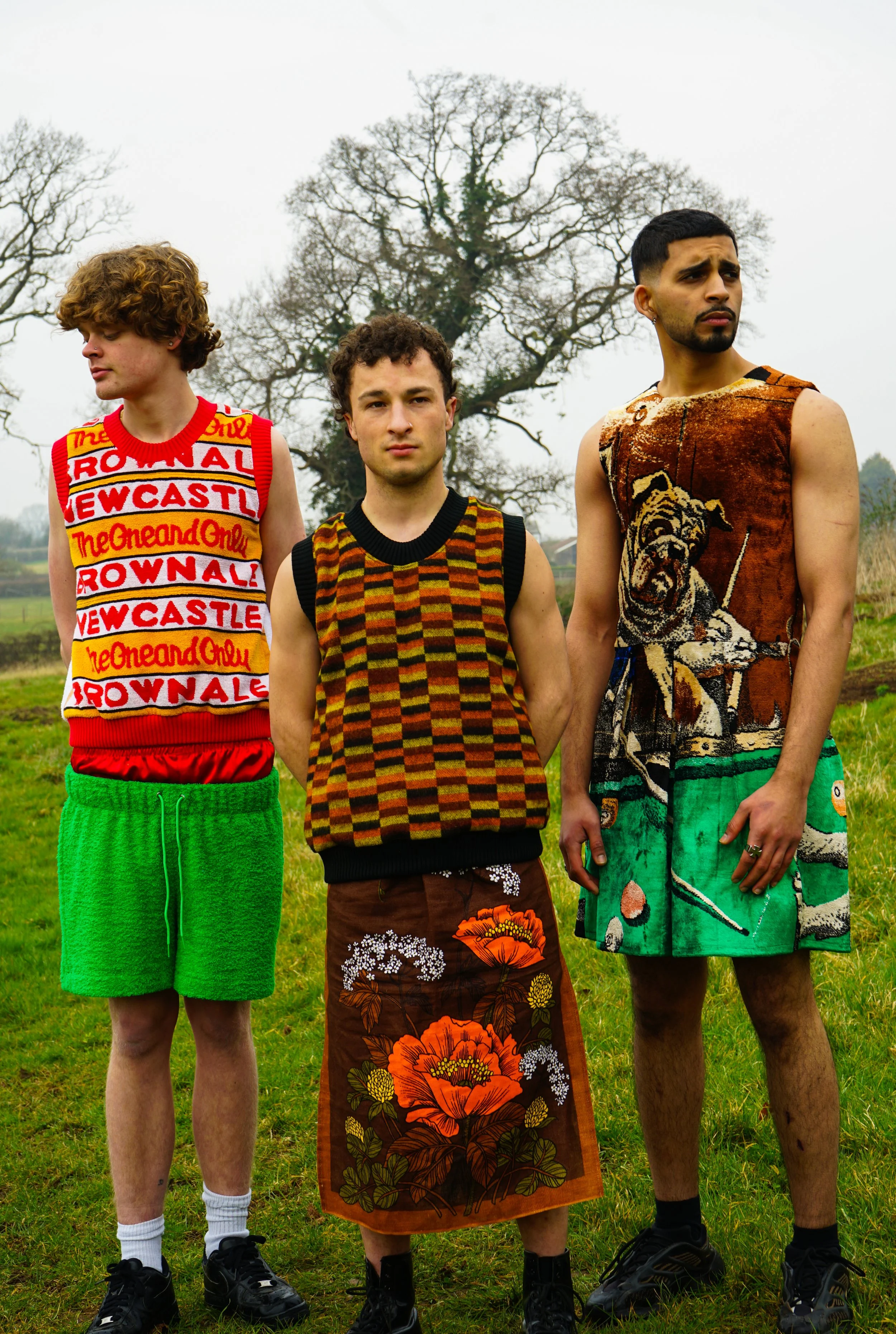 Three young men standing outdoors on a grassy field with leafless trees in the background, wearing colorful, eclectic clothing.