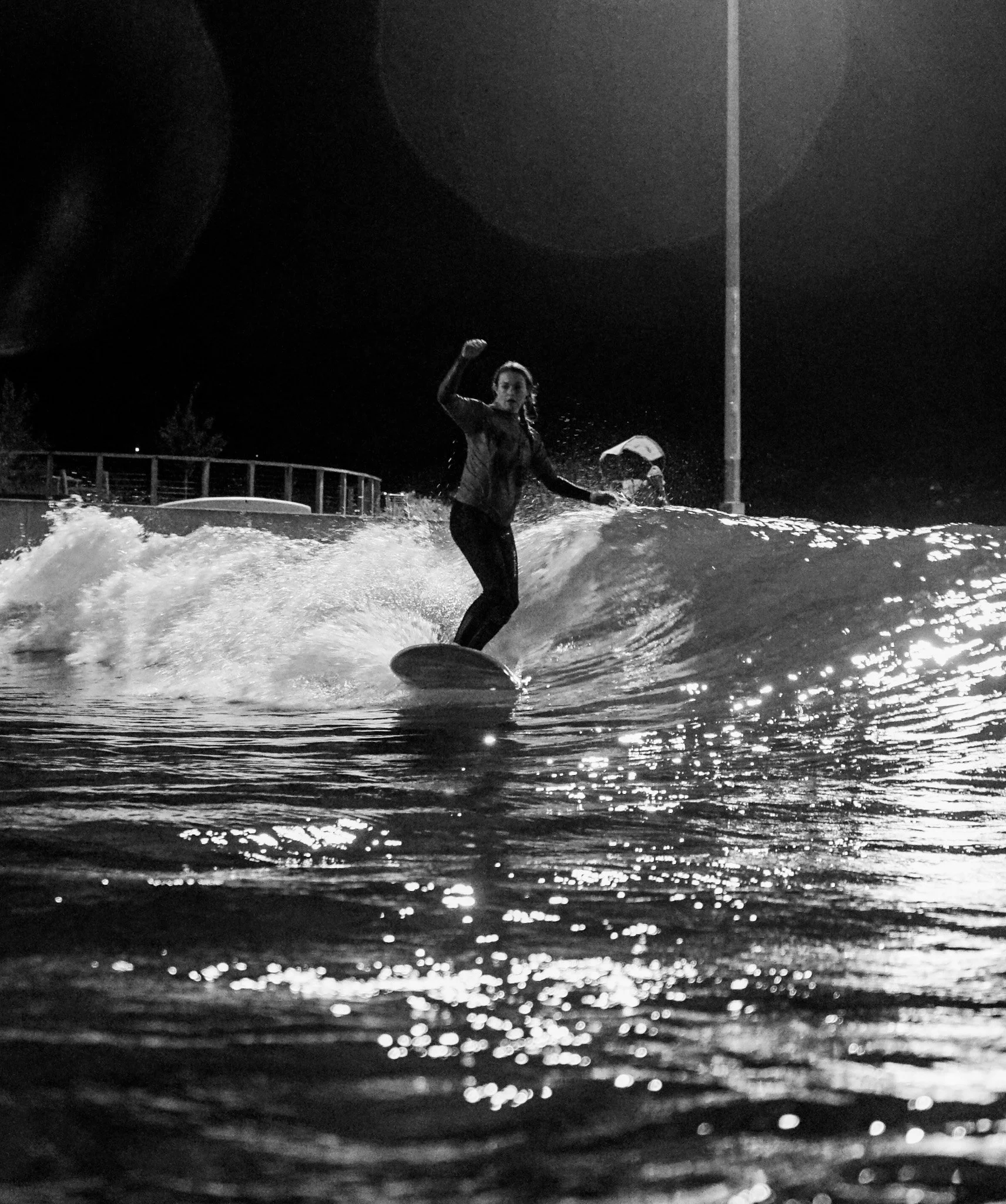 A woman surfing on a wave at night, wet from the water, with her right fist raised, in a scene illuminated by reflections on the water surface and a streetlamp in the background.
