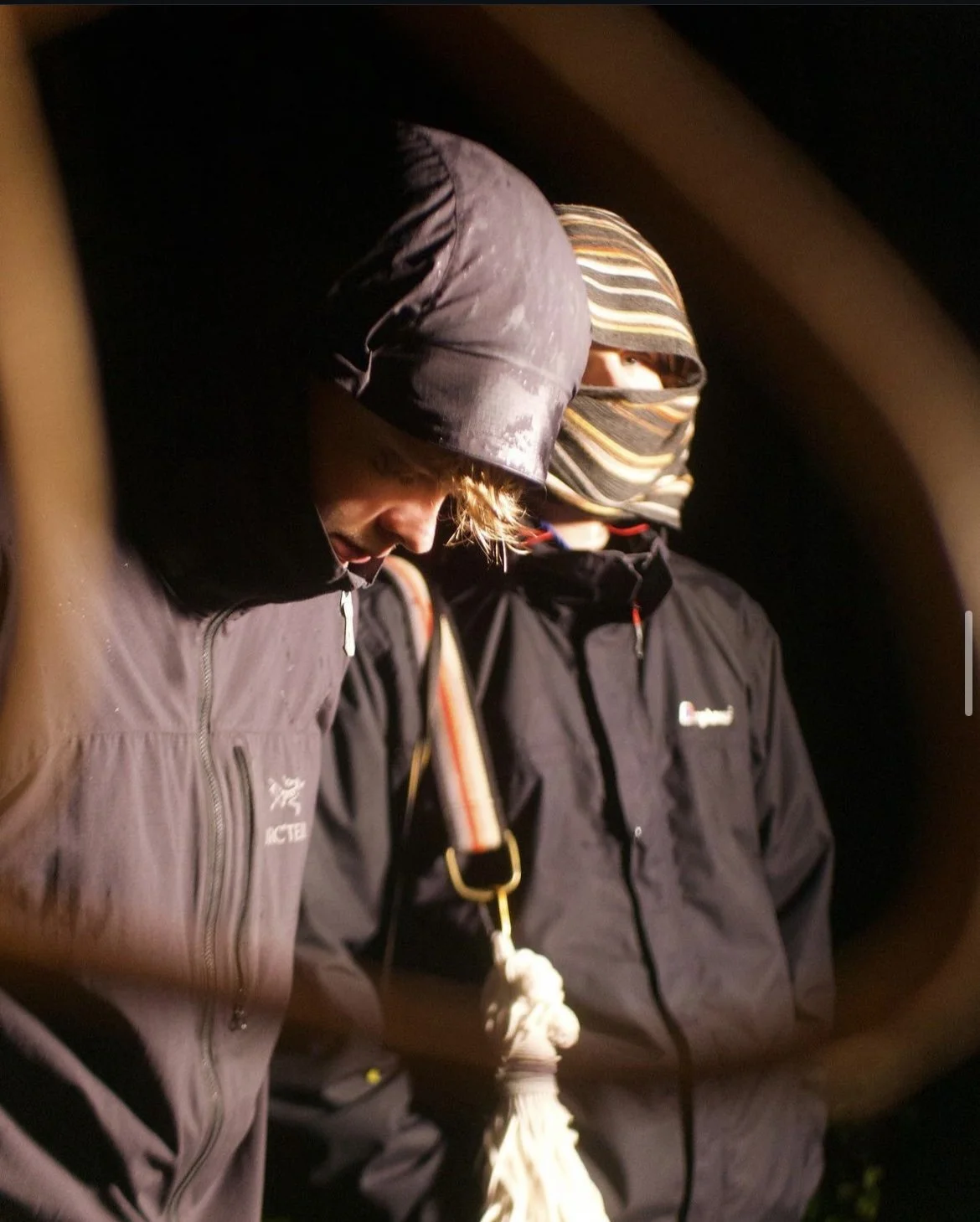 Two individuals wearing jackets, beanies, and face coverings, standing closely at night. The person on the left has a black beanie and a hood, looking down. The person on the right has a striped beanie and a face covering, looking slightly downward. 