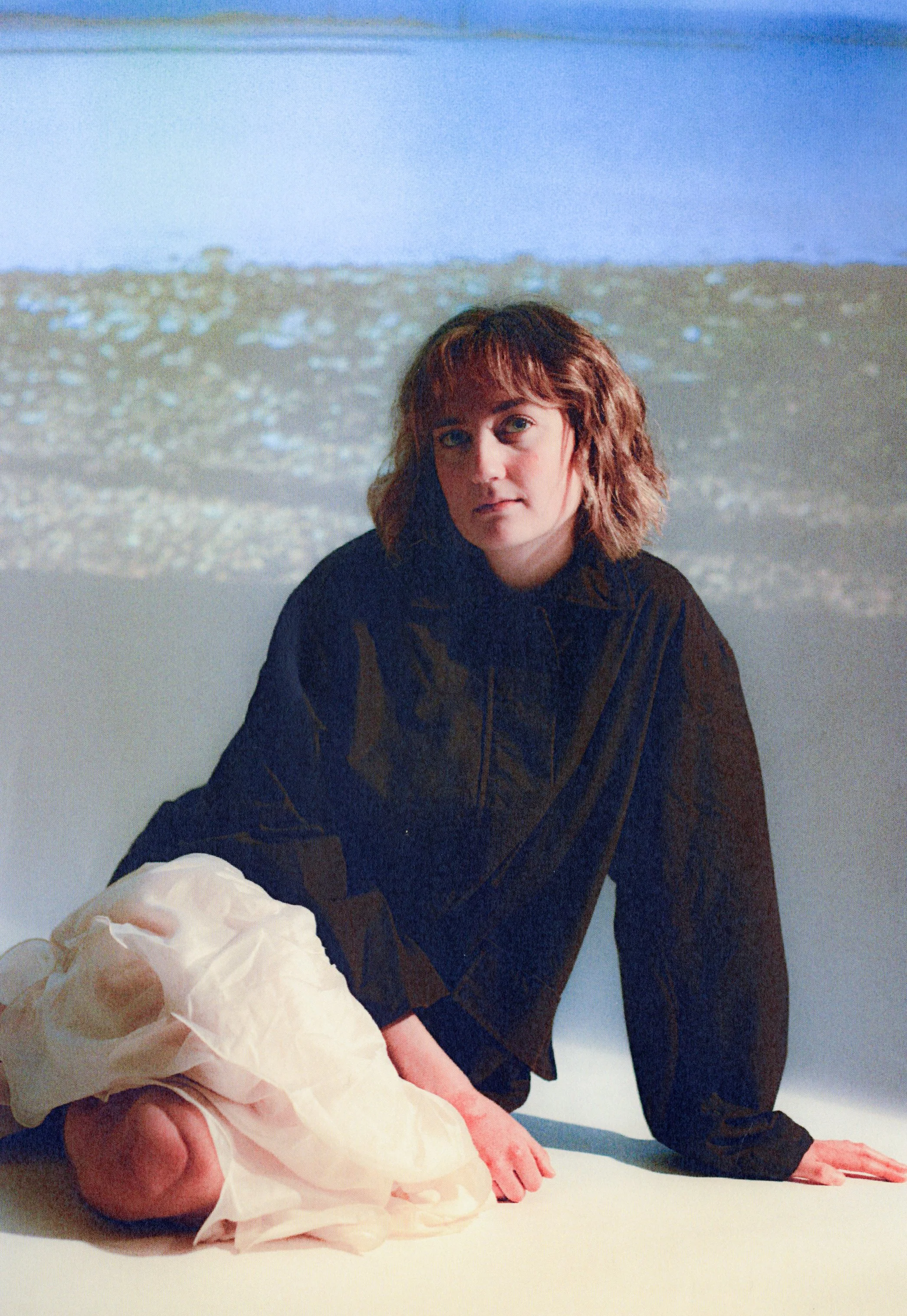 A woman with short wavy hair and fair skin is sitting on the floor, wearing a black long-sleeved top. She is leaning forward on her hands. A hand holding crumpled white tissue paper is extended towards her from the left side of the image. In the back