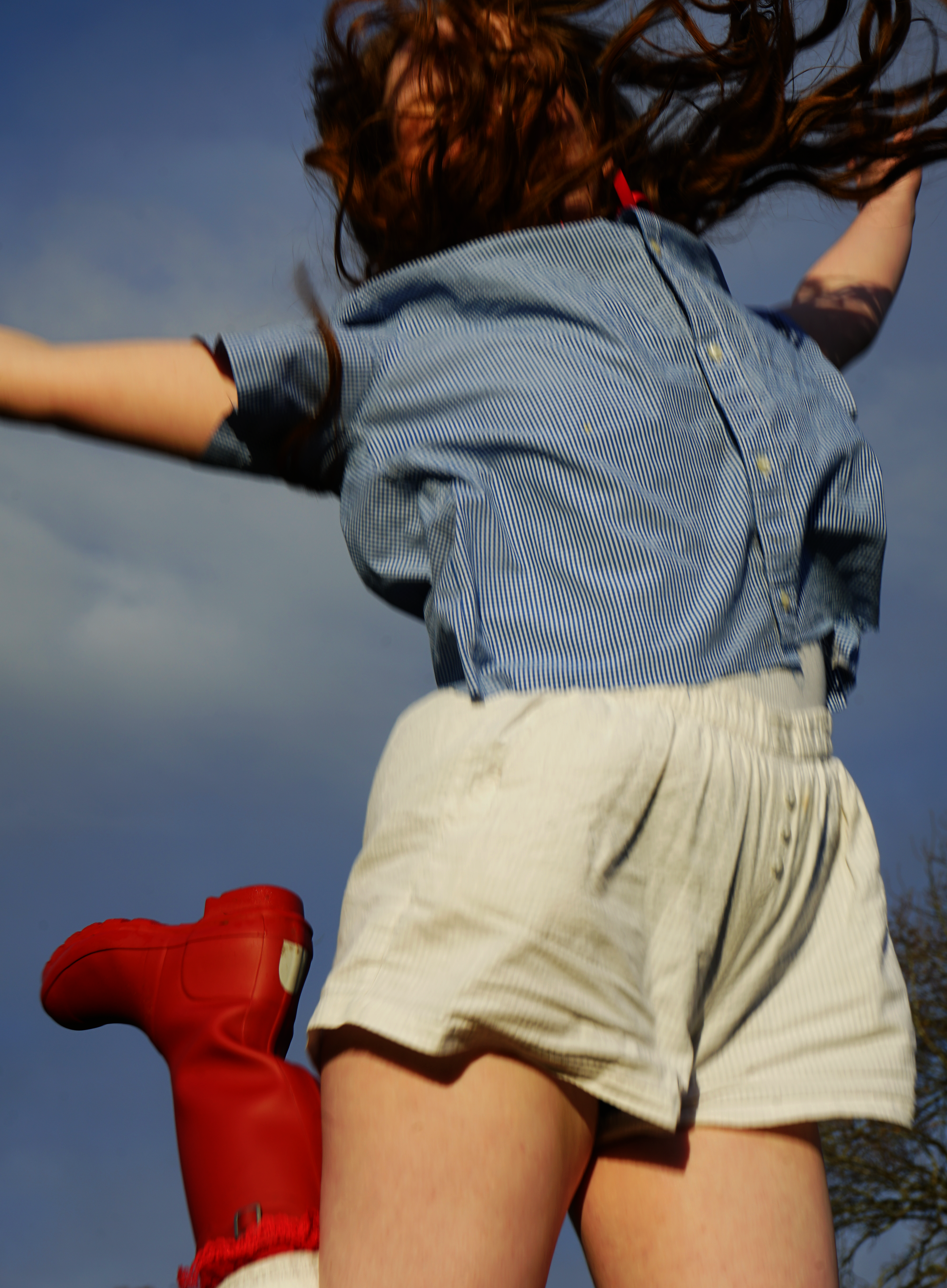 Close-up of two people from behind jumping outdoors on a cloudy day, one in a blue striped shirt and beige shorts, the other in red shorts.