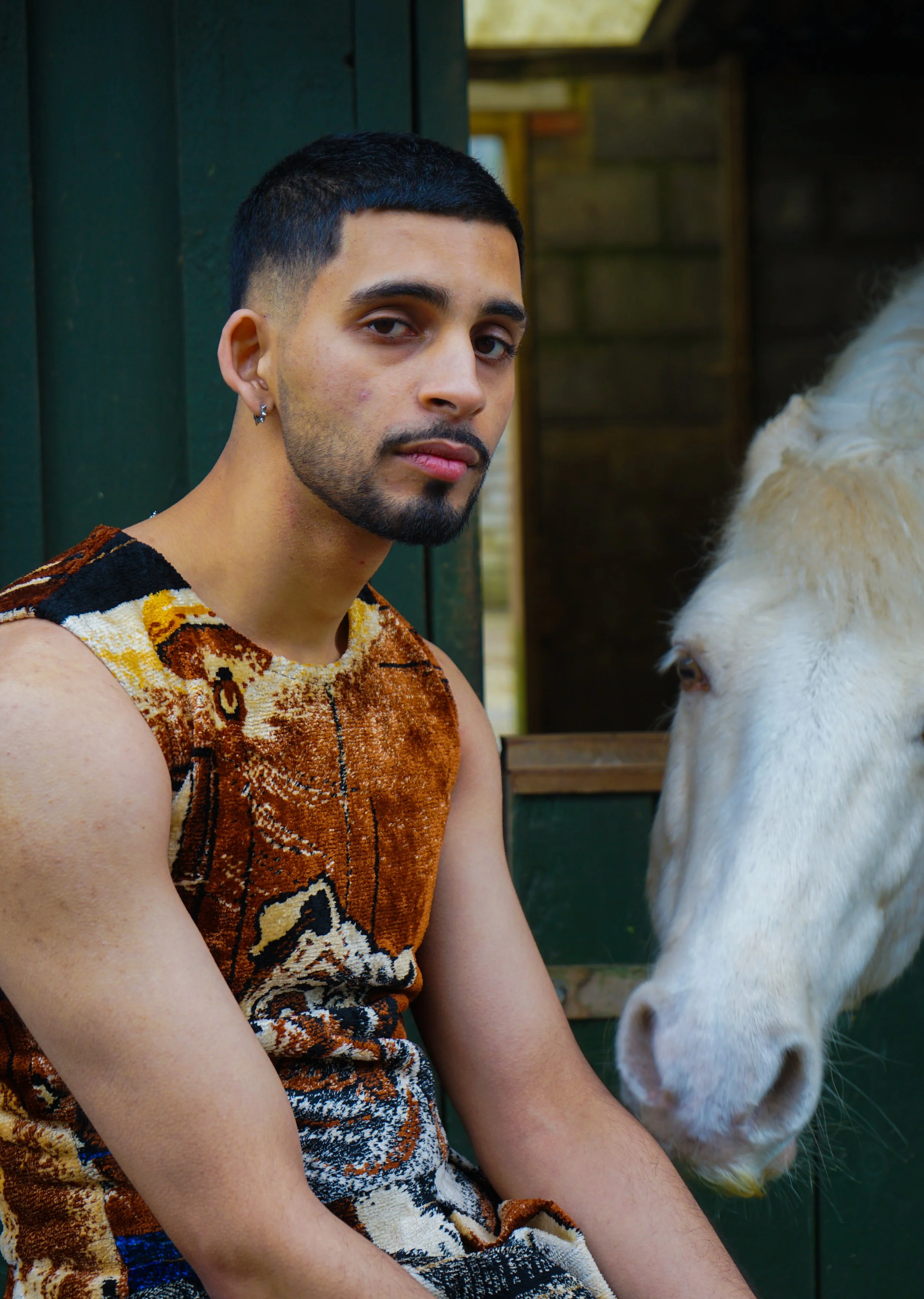 A young man with short black hair, a beard, and earrings sitting next to a white pony or horse, both outside near a green wooden structure.