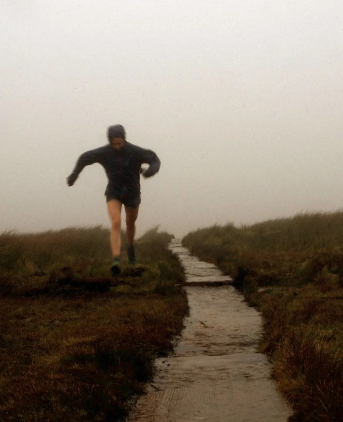 A person running on a narrow dirt trail through a foggy, grassy landscape.