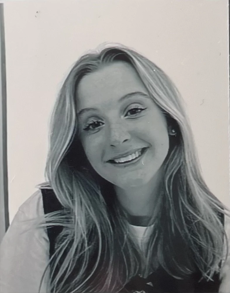 Black and white photograph of a smiling young woman with long wavy hair, wearing a light-colored shirt and dark overalls.