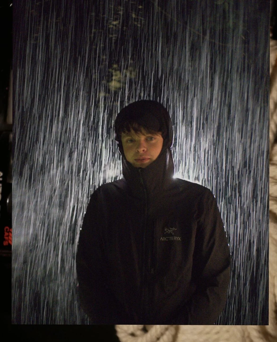 A person wearing a black Arc'teryx jacket standing under a heavy rainfall at night, with rain streaks illuminated by a light source behind them.