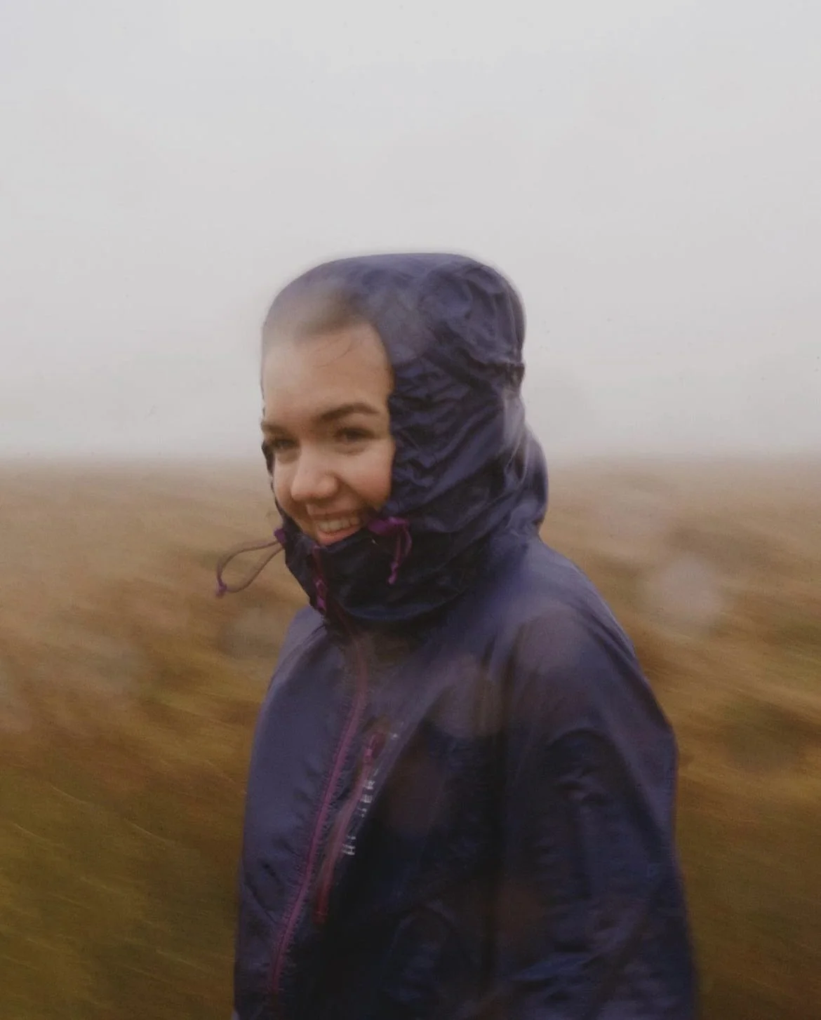 A woman outdoors in a field during foggy weather, wearing a dark rain jacket with the hood up, smiling.