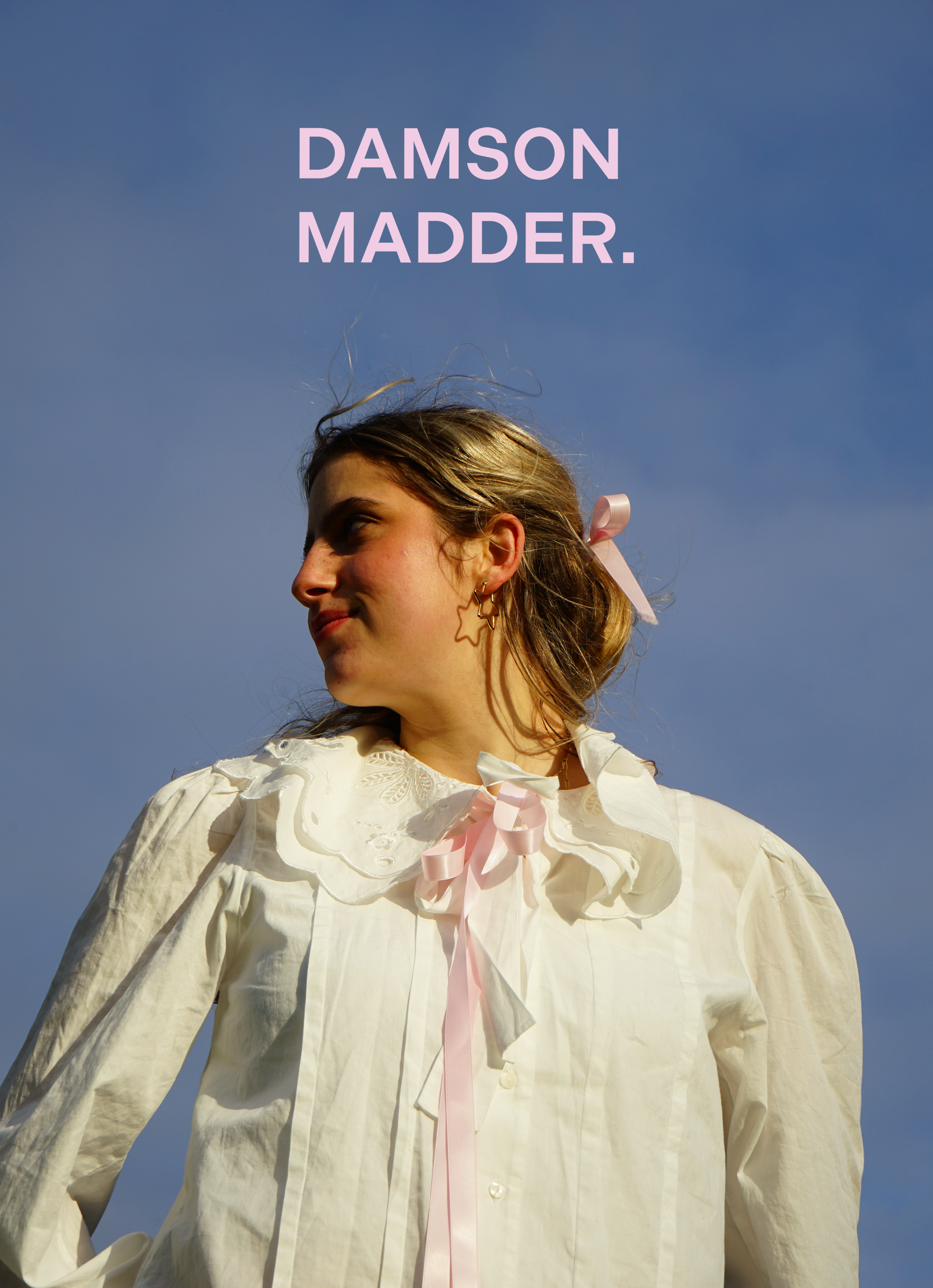 A young woman with wavy hair tied with a pink ribbon, wearing a vintage cream-colored blouse with a large collar and pink ribbon tie, standing outdoors against a blue sky.