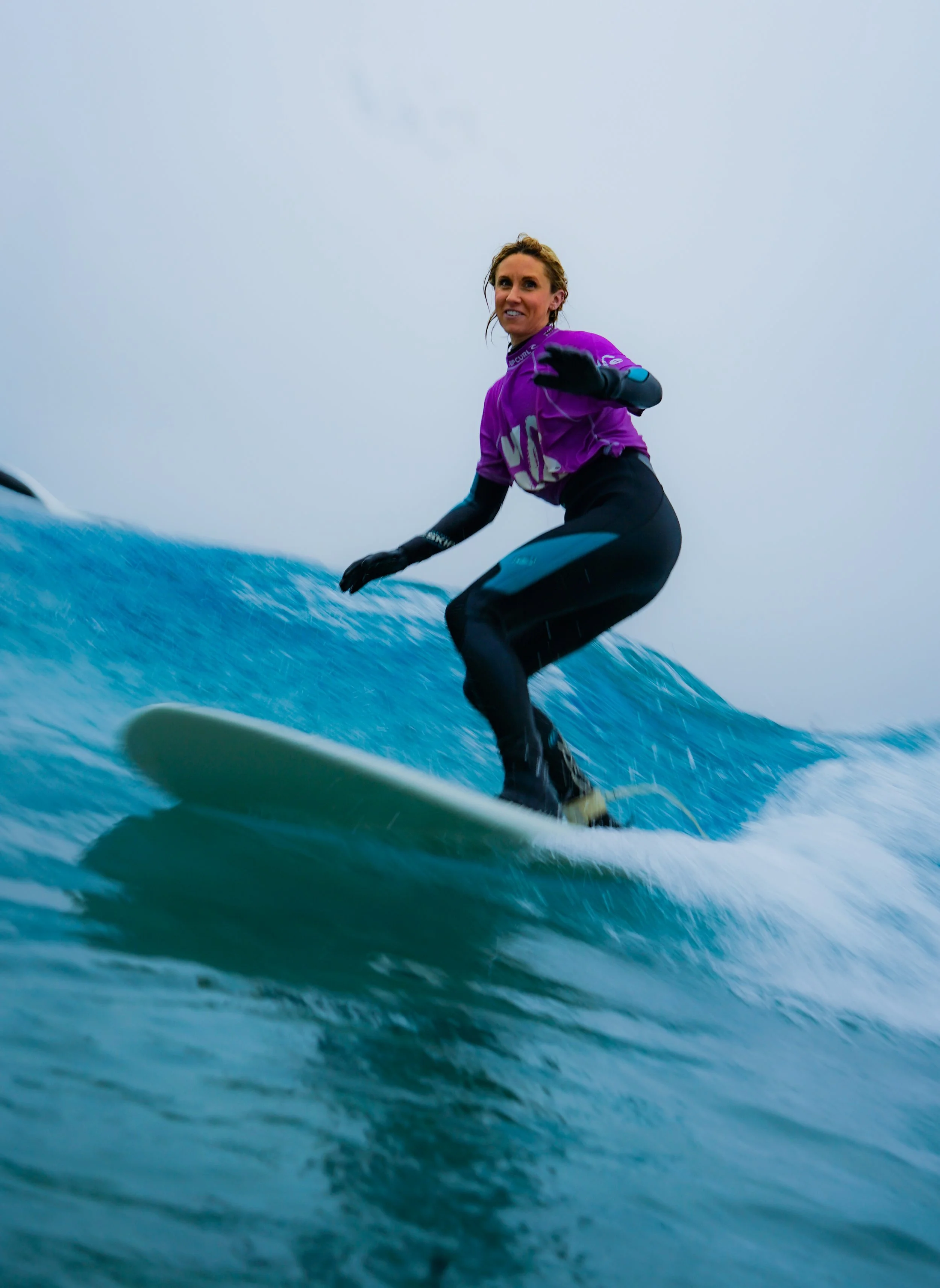 A woman in a purple shirt and black pants surfing on a blue wave during cloudy weather.