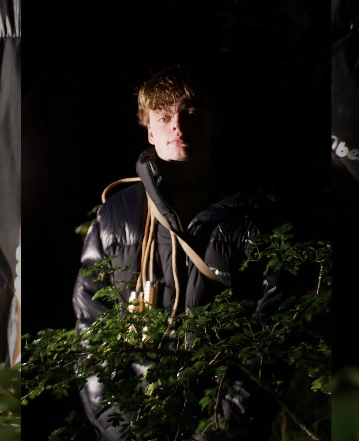 A person with curly hair wearing a black jacket with a tan cord strap, surrounded by green foliage in a dark environment.