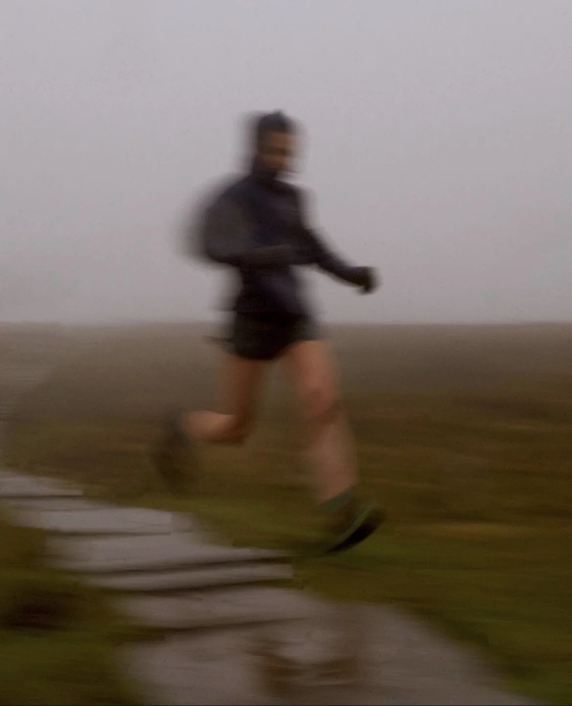 A person running outdoors in a foggy environment, wearing a dark jacket, shorts, and running shoes, with a blurred background and stacked white items on the ground.