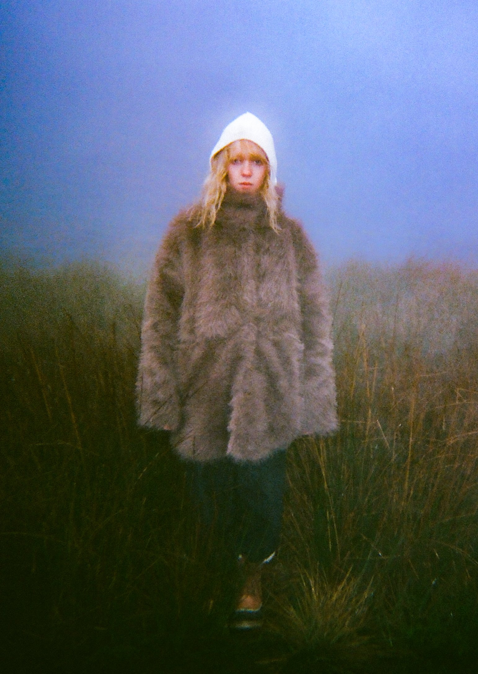 A person standing outdoors in a foggy or misty environment, wearing a white beanie and a thick brown fuzzy coat, with long wavy hair and partially visible shoes, surrounded by tall grass.
