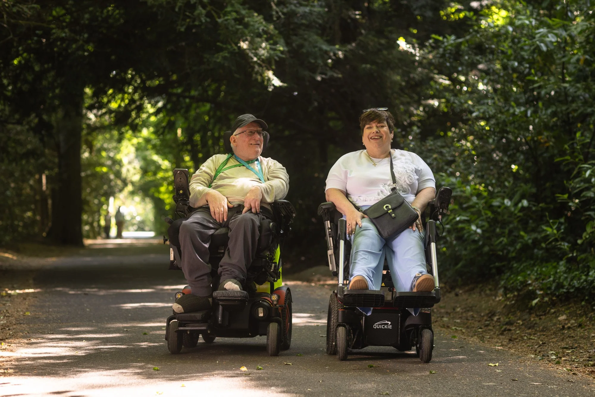 A man and a woman, both in wheelchairs, sitting outdoors on a tree-lined path, smiling and enjoying conversation.