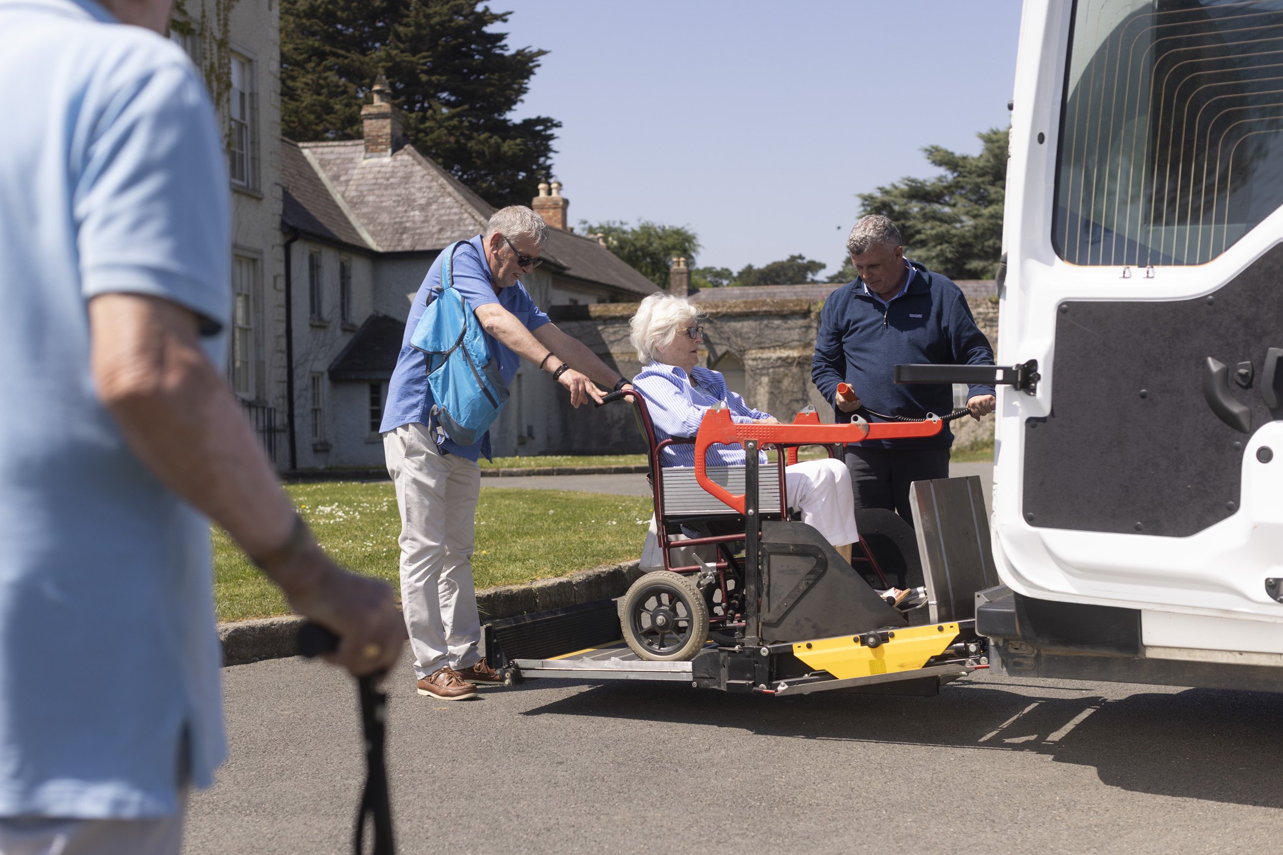 An elderly woman in a wheelchair being assisted into a vehicle by two men, one pushing the wheelchair and the other holding a device, on a street with houses and trees in the background.