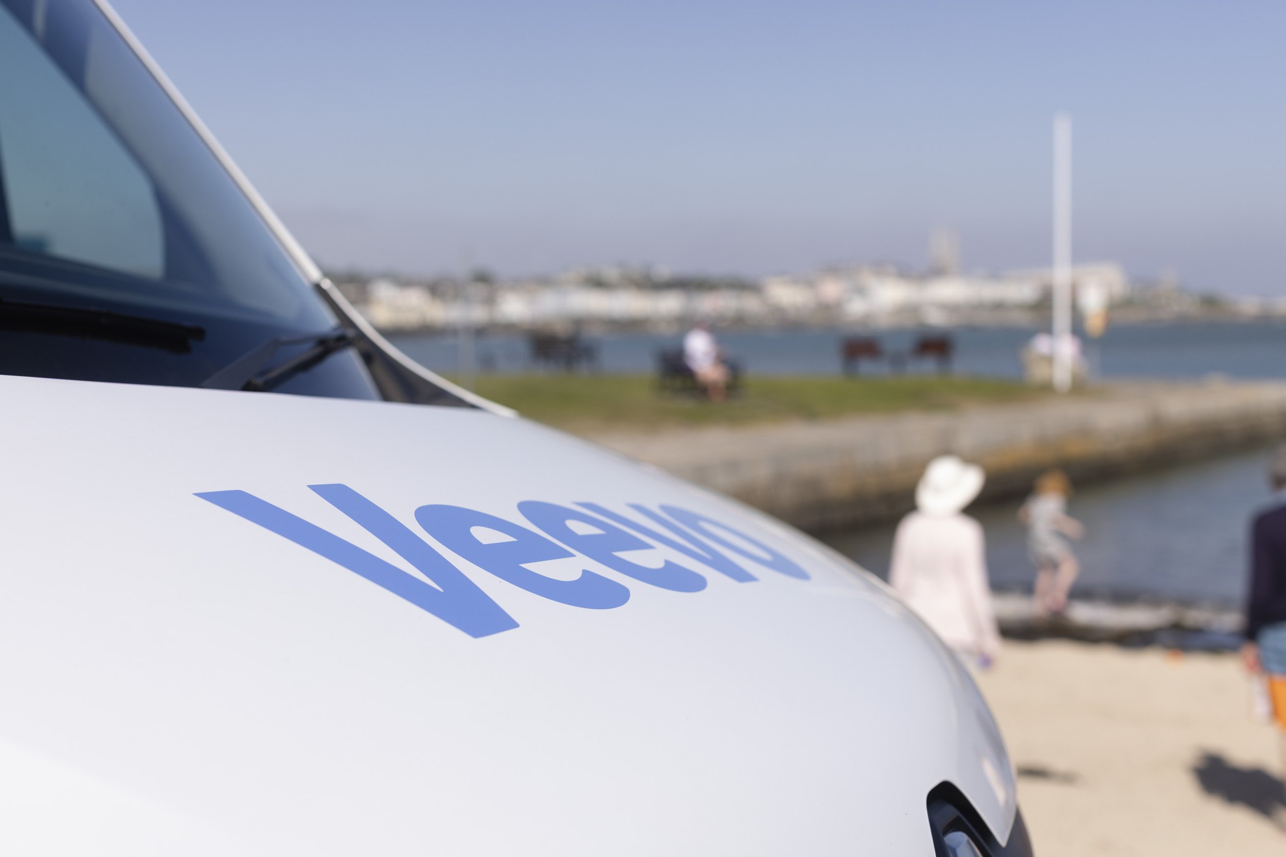 Close-up of a white car with the blue text 'Uber' visible, parked near a waterfront with people sitting on benches and a bridge in the background on a clear day.