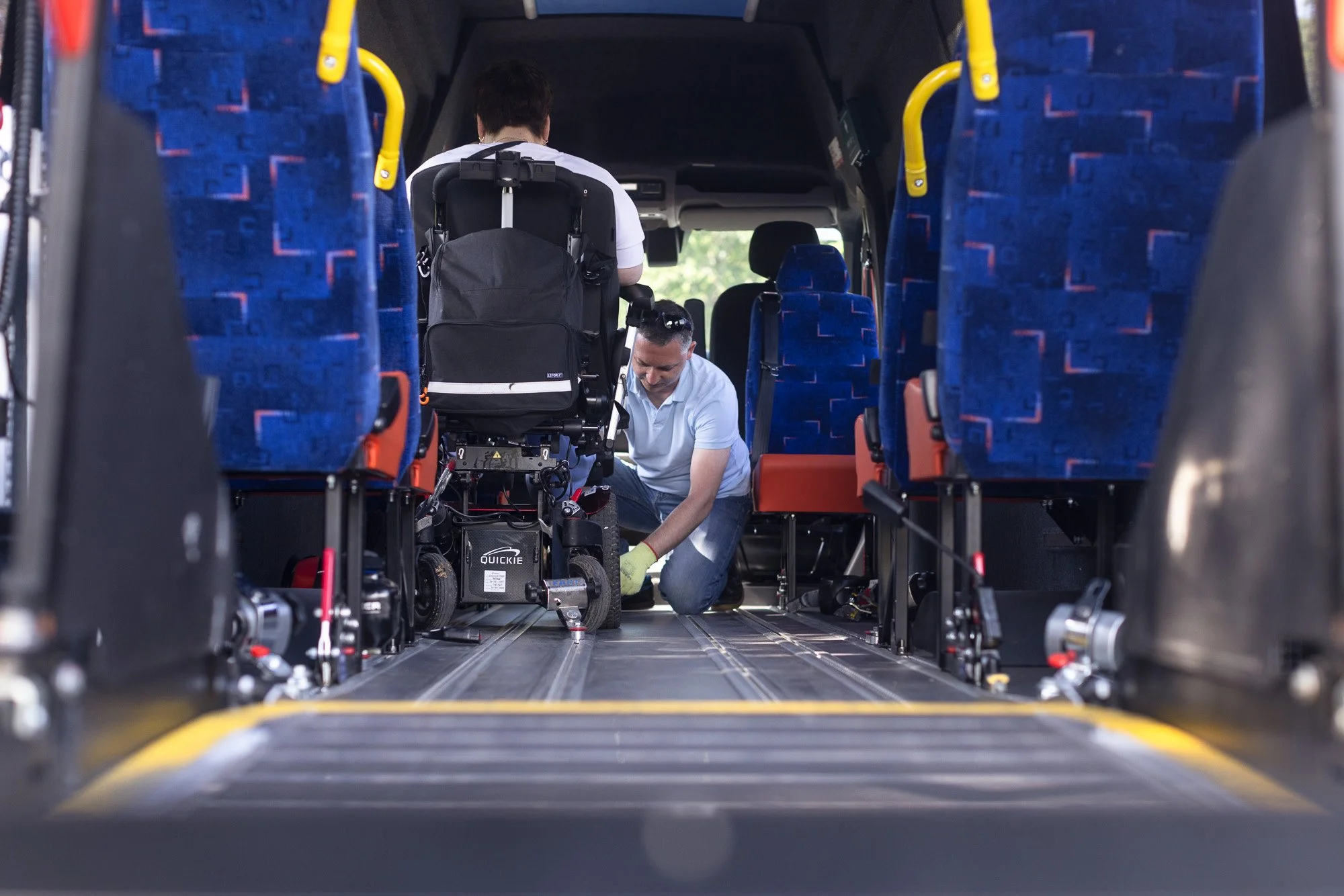 Two men are inside a bus, working on equipment. One man is kneeling on the floor and the other is standing, with a stroller on his back. The bus seats are seen on either side, with blue upholstery and orange accents.