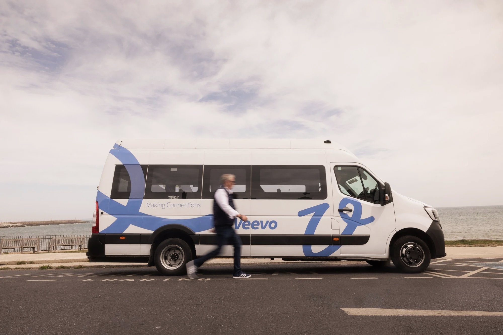 A white van with blue graphics and the word 'Veevo' parked beside a concrete promenade. A person is walking in front of the van, and the sky is cloudy.