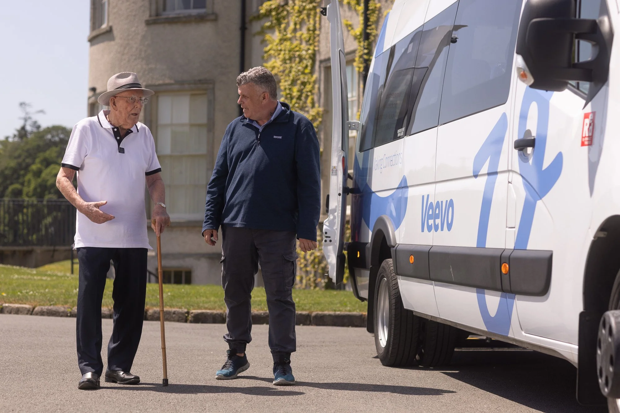 An elderly man with a walking cane talking to a middle-aged man in front of a large delivery van with 'Volvo' branding. The elderly man is wearing a hat and white polo shirt, while the middle-aged man is dressed in casual dark clothing and sneakers. They are outdoors in front of a residential building with some greenery.